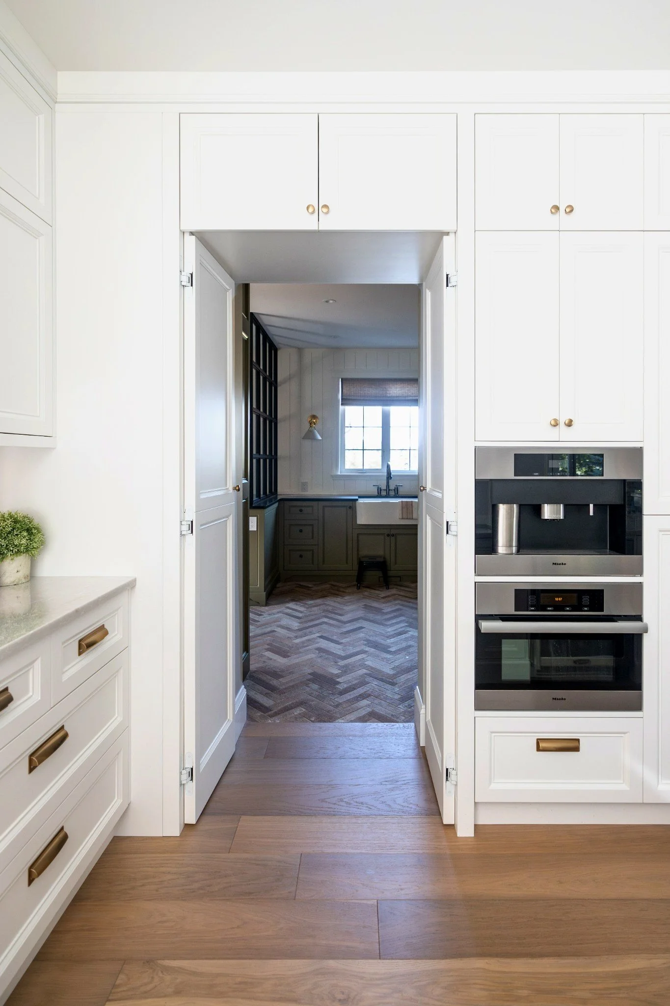 View from a kitchen into a small but inviting breakfast nook with a window and a sink, featuring earth-tone cabinets and chevron-patterned wooden flooring.