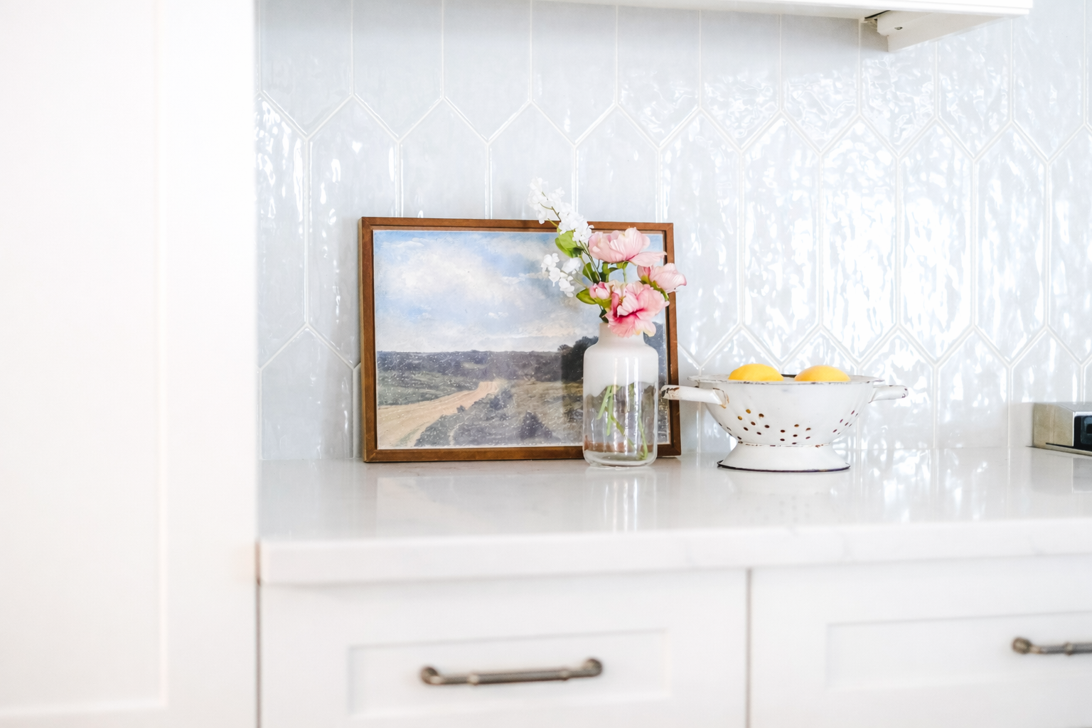 Decorative kitchen countertop with a framed landscape painting, a white vase with pink and white flowers, and a white colander with lemons against a glossy hexagonal tiled wall.