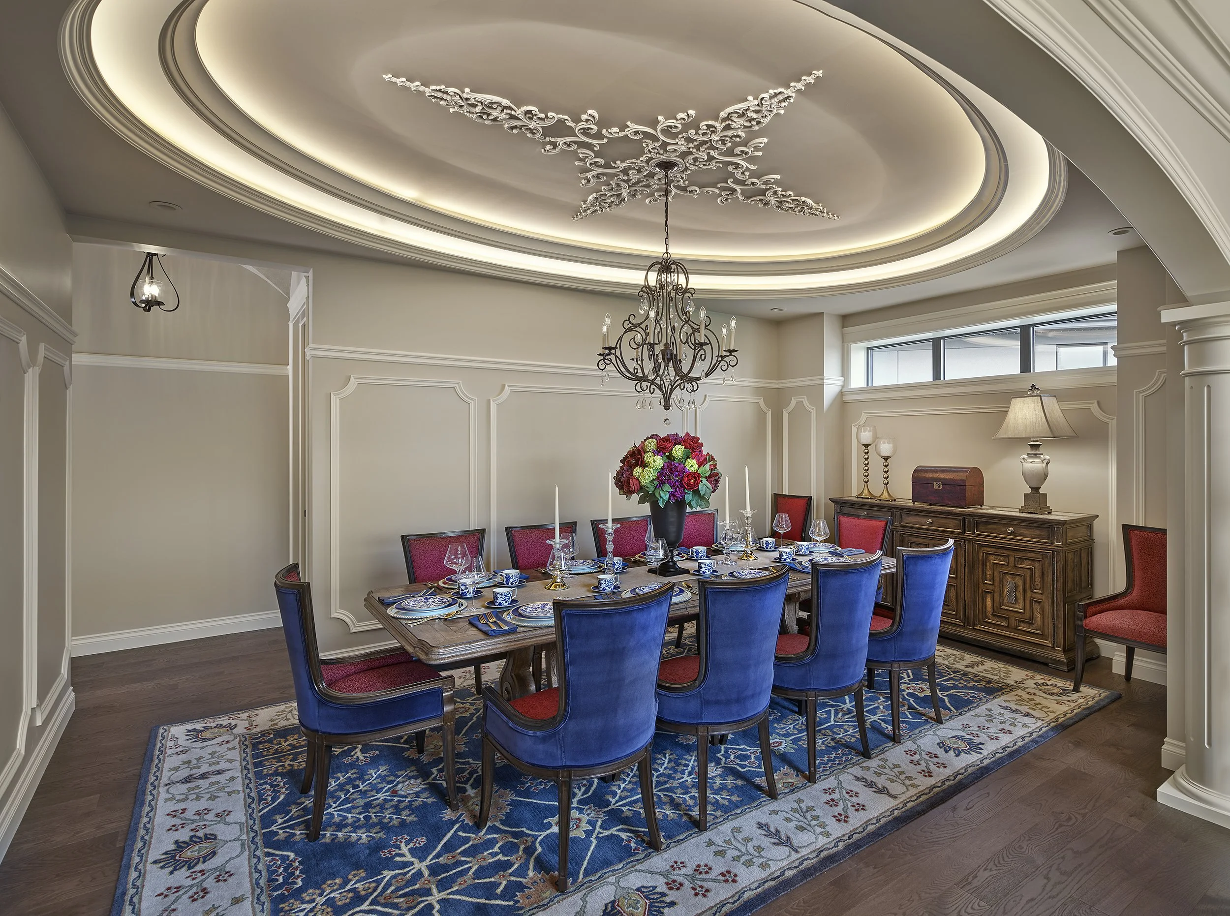 Elegant dining room with a long wooden table, surrounded by blue and red upholstered chairs, a floral centerpiece, and decorative wall panels, with a chandelier hanging from a detailed ceiling.