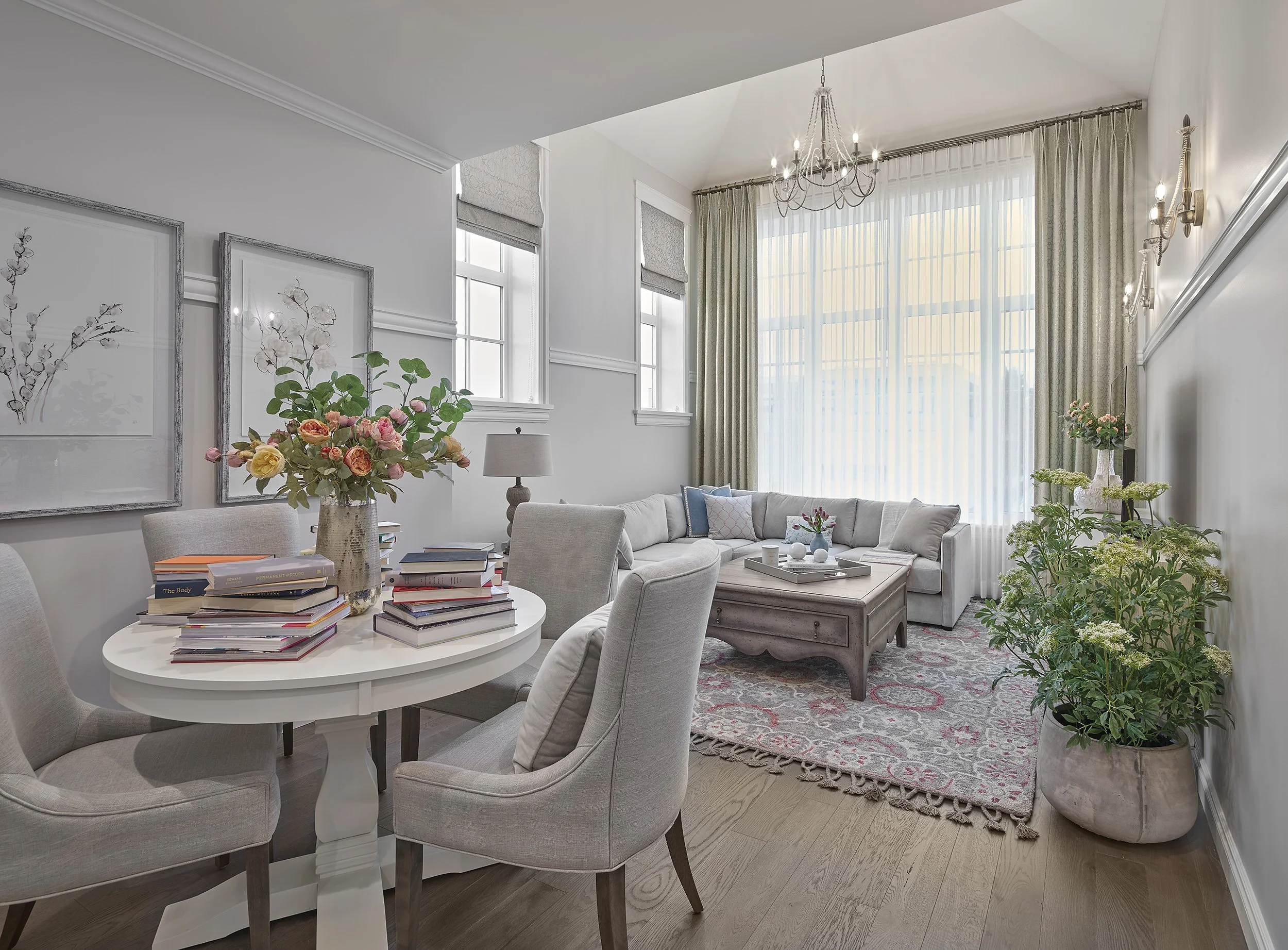 Bright living room with white walls, large windows with sheer curtains, a chandelier, light-colored sofa, wooden coffee table with decorative items, floral area rug, potted plants, and a round dining table with stacked books and a vase of flowers.
