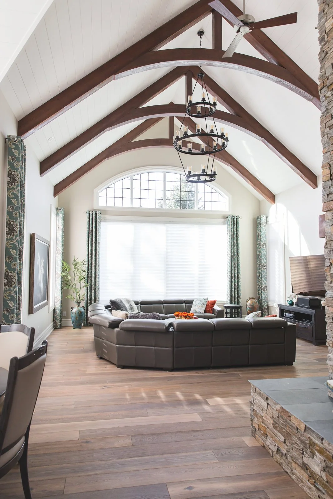 Living room with vaulted ceiling, wooden beams, large window with curtains, round leather sofa, chandelier, ceiling fan, fireplace with stone surround, and wooden flooring.