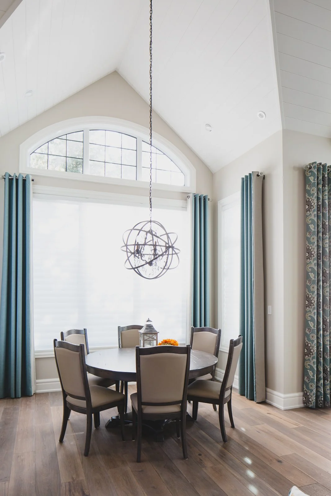A dining room with a round table surrounded by six beige upholstered chairs, a metal lantern and a flower on the table, large arched window and tall curtains, and a modern globe chandelier hanging from a vaulted ceiling.