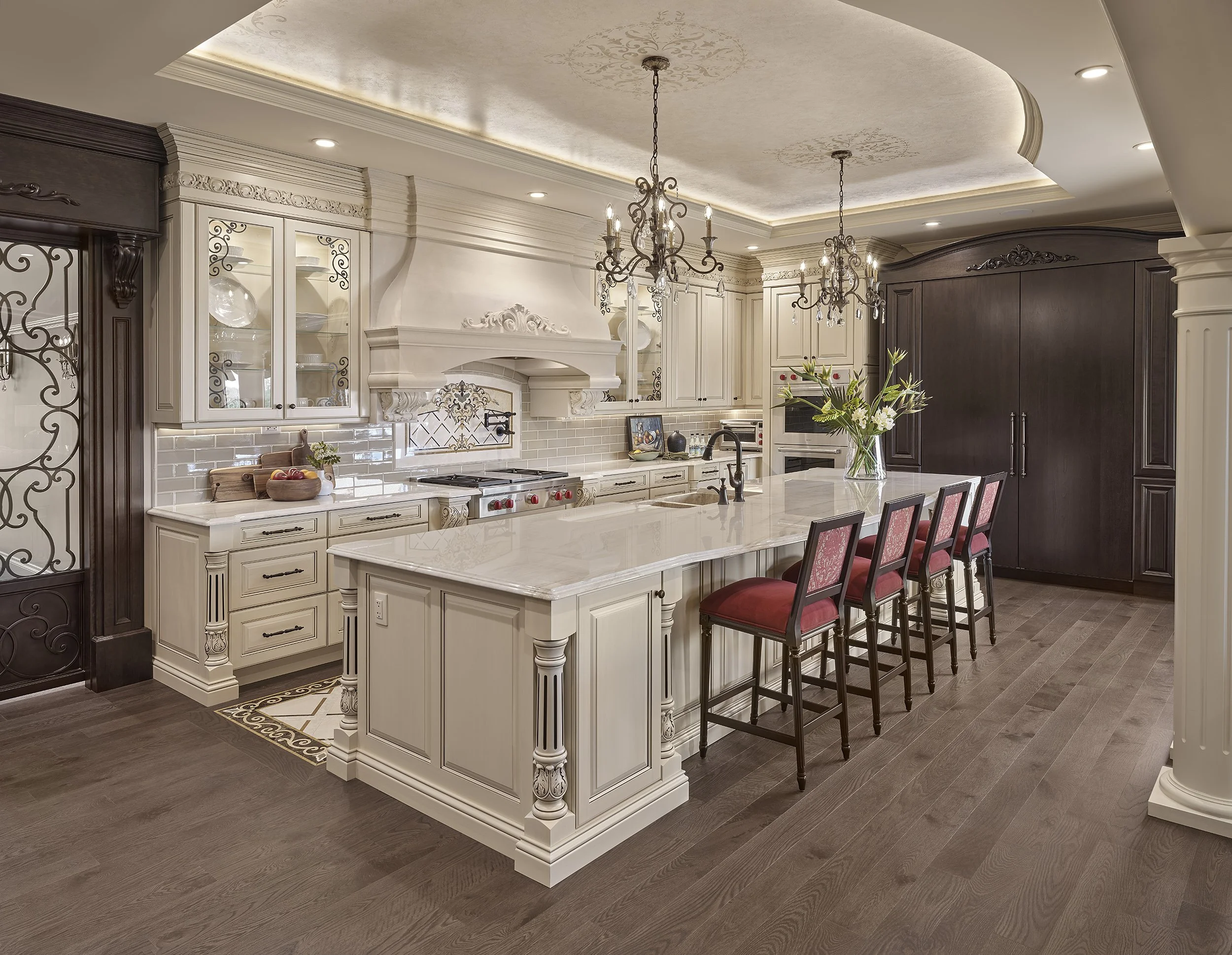Elegant kitchen with white cabinets, a large marble island with five red cushioned chairs, chandelier lighting, dark wood flooring, and a mix of white and dark cabinetry.