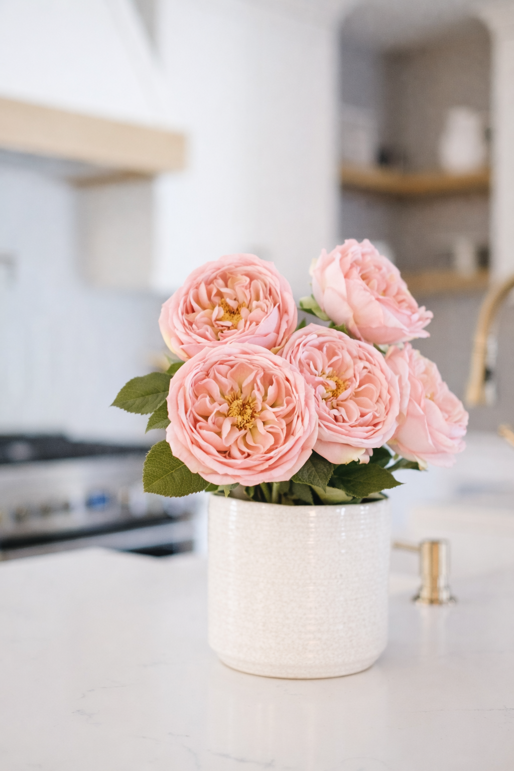 A bouquet of pink roses in a textured white ceramic vase on a white countertop in a modern kitchen.