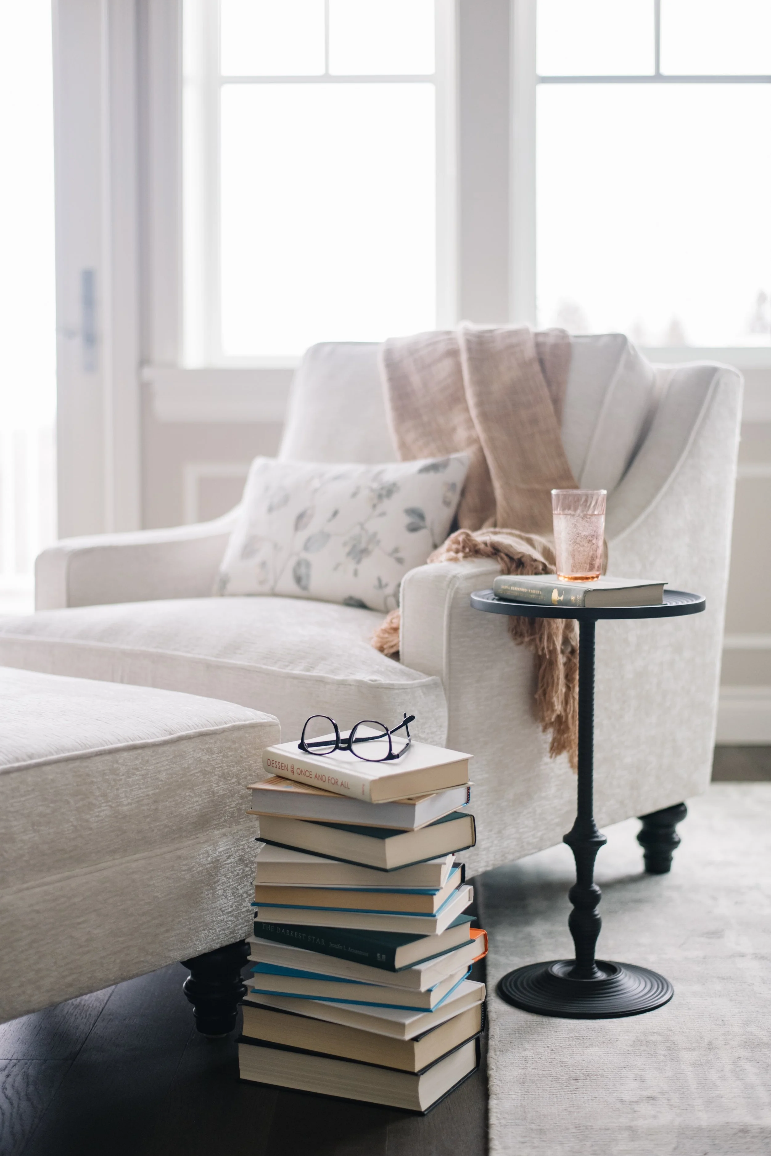 Cozy living room corner with a white armchair, standing on a light-colored rug, next to a tall stack of books with glasses on top. There is a small black side table with a glass of rosé wine and a closed book on it, in front of a window letting in na