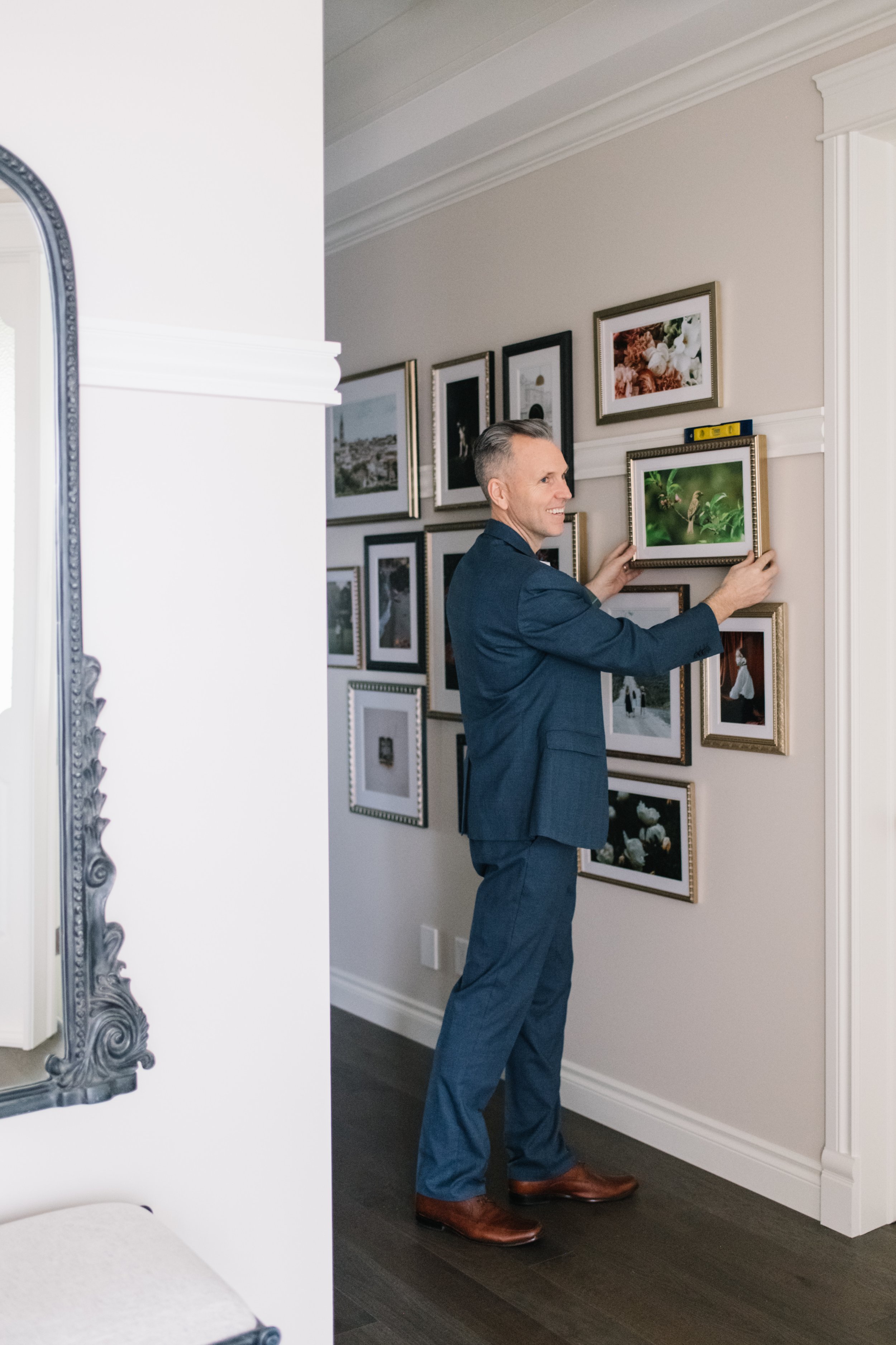 A man in a blue suit adjusting a framed photograph on a gallery wall inside a home, with other framed pictures behind him.
