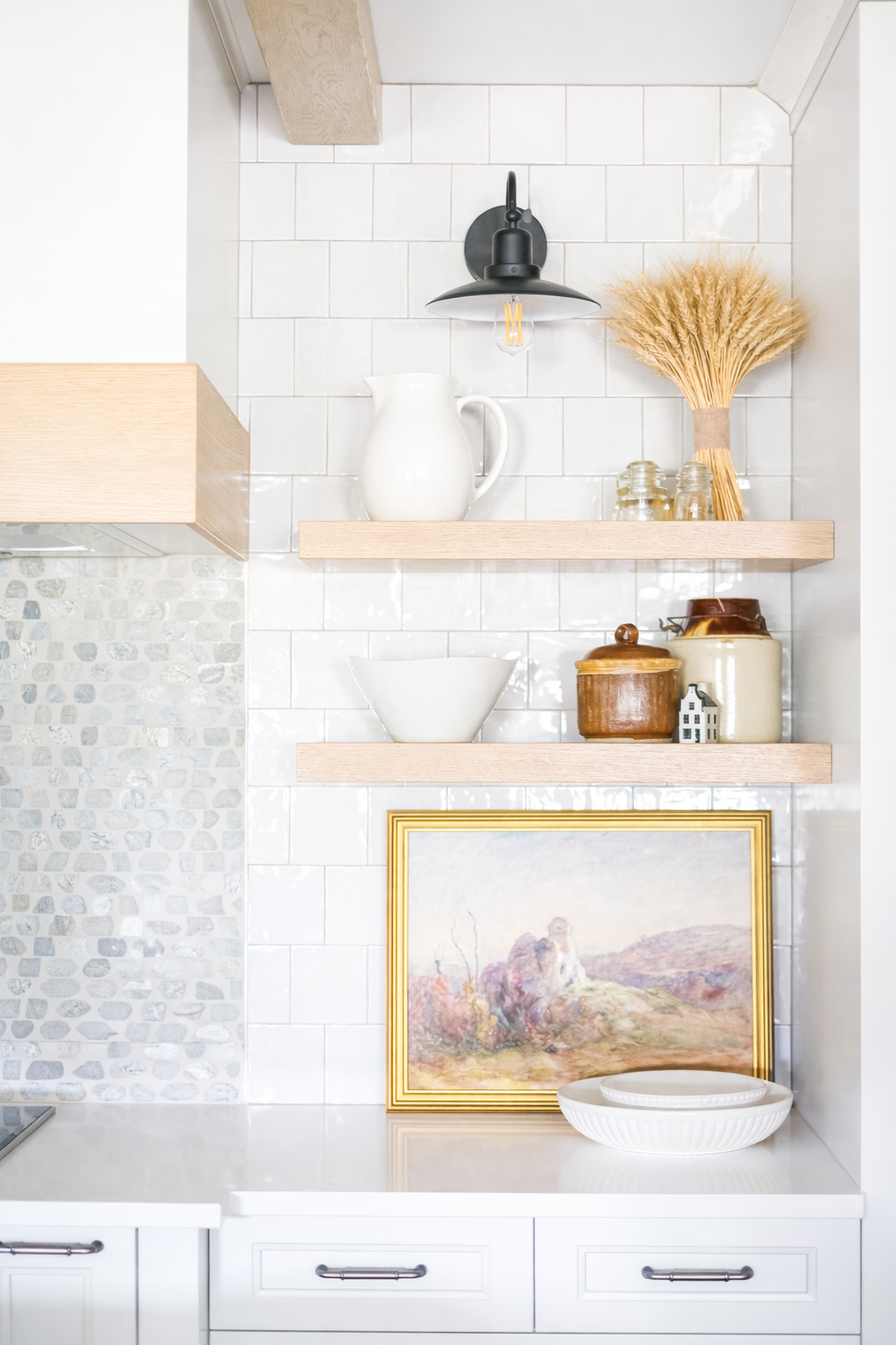 Decorative kitchen with white tile backsplash and light wood open shelves holding a white pitcher, dried wheat bouquet, jars, a white bowl, ceramics, a framed painting, and white dishes.