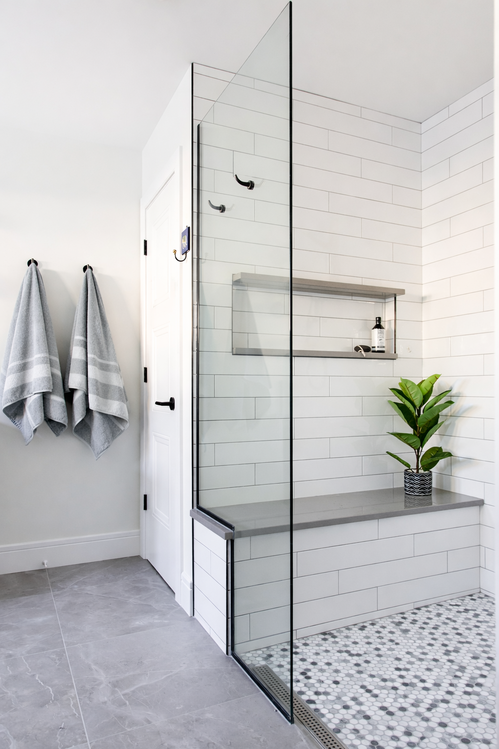 Modern bathroom with a walk-in shower enclosed by a glass wall and door, white subway tile walls, a gray bench, a potted green plant, and two hanging gray towels.