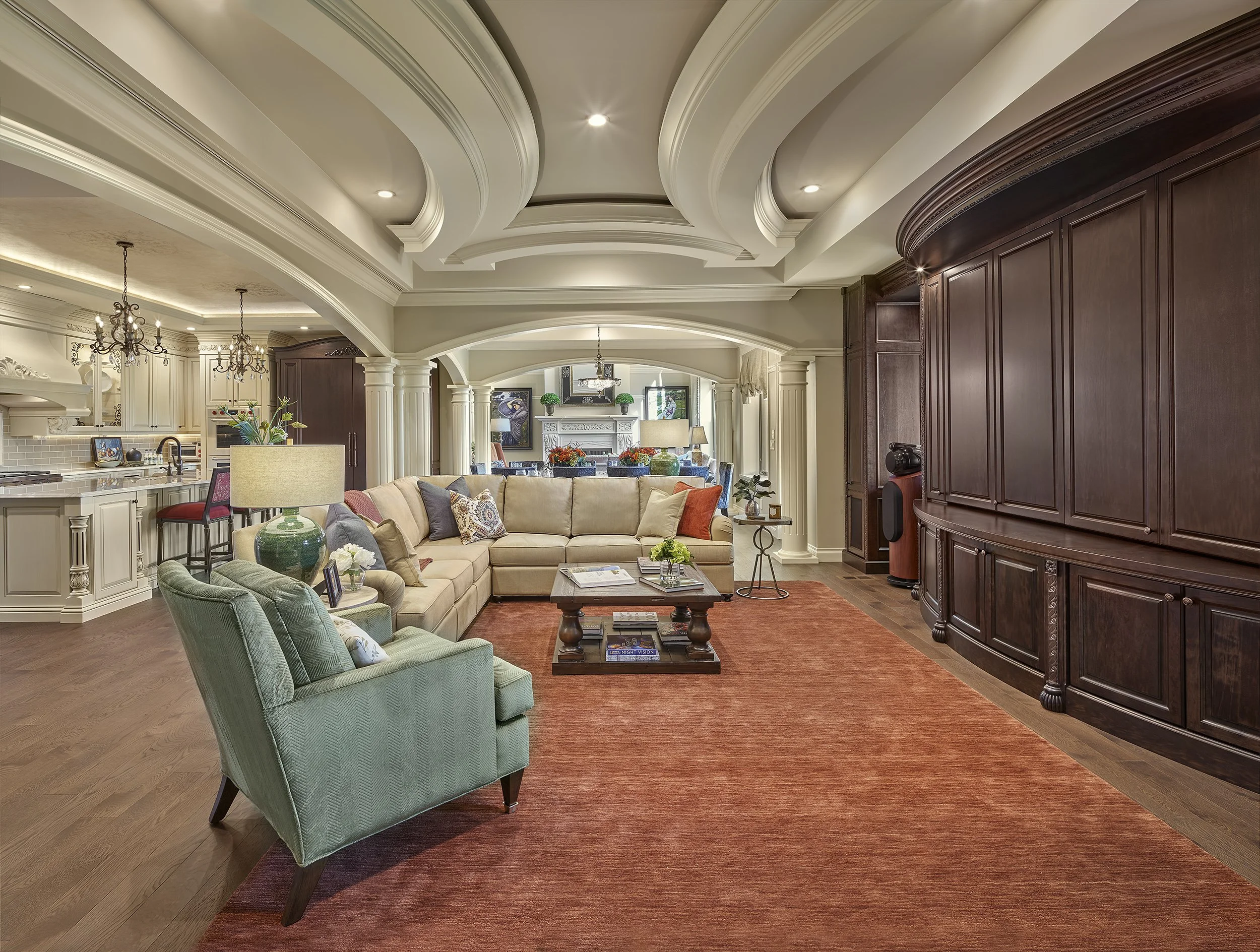 A spacious, elegantly decorated living room with a large beige sectional sofa, a green armchair, a wooden coffee table, and a wooden built-in wall unit. The room features crown molding, columns, and a red area rug on hardwood floors. In the backgroun
