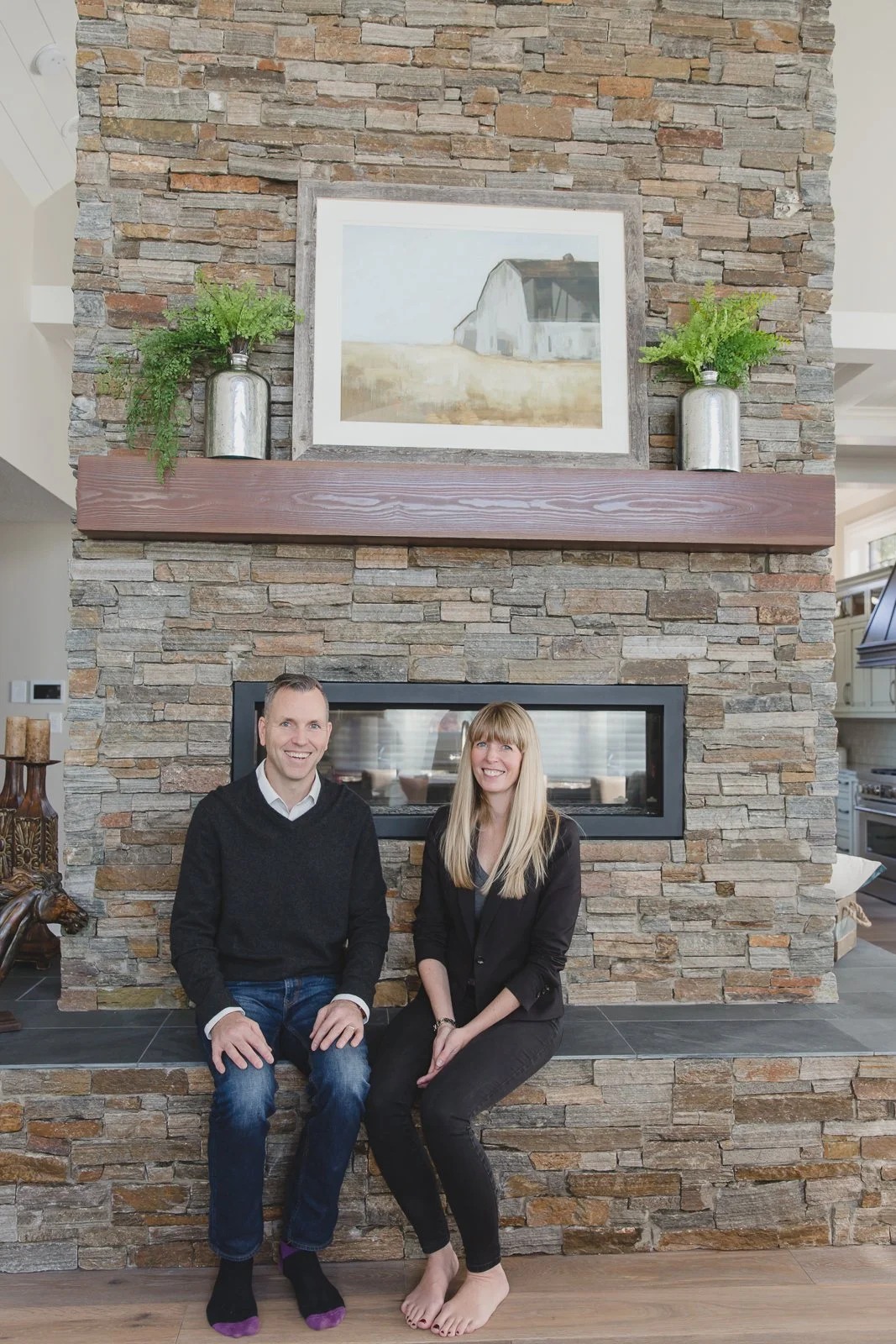A man and a woman sit on a stone hearth in front of a fireplace, smiling. The background features a stone wall with a framed picture of a barn and two silver vases with green foliage on a wooden mantel.