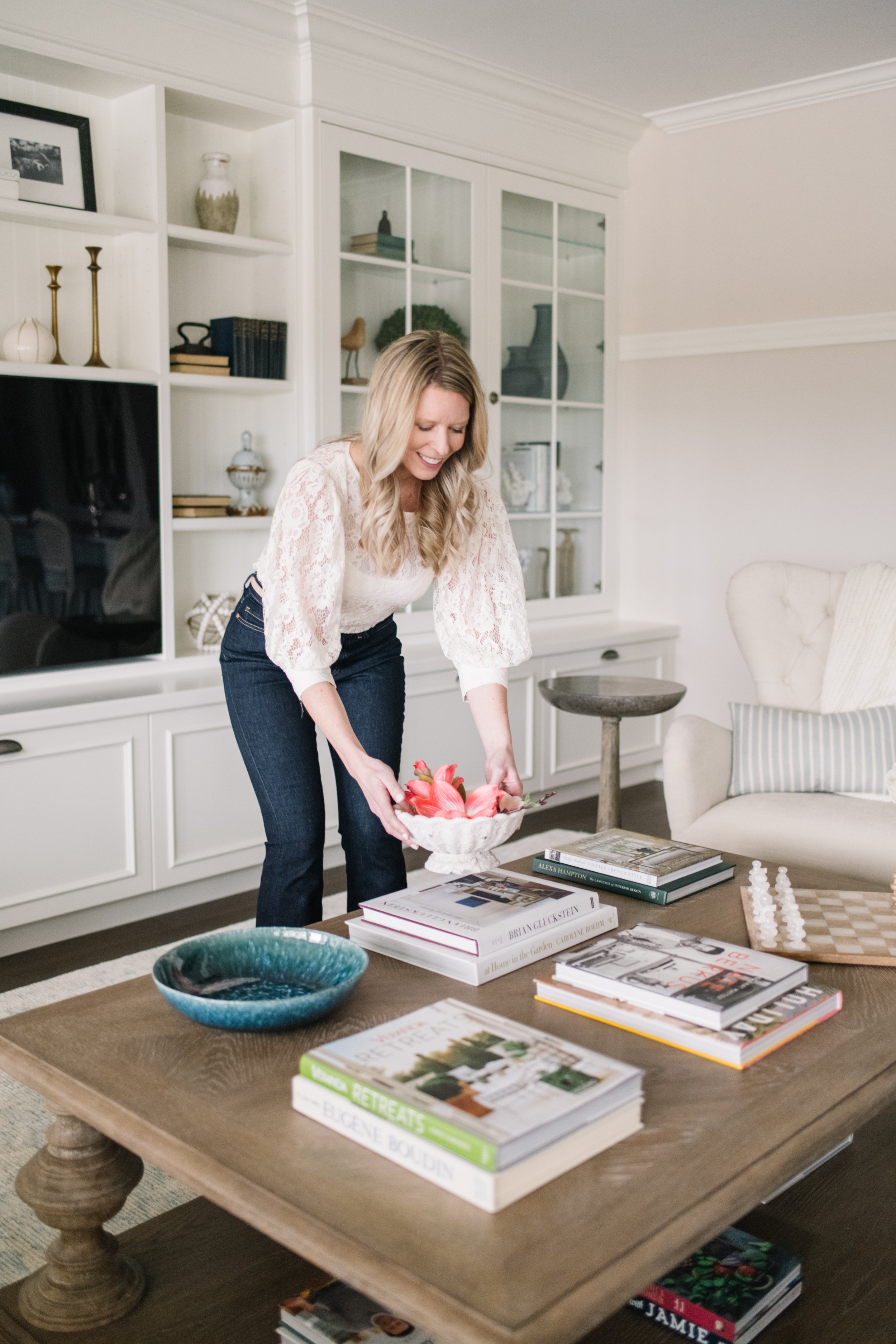 A woman with blonde hair wearing a white lace top and dark jeans is smiling while placing a bowl with pink flowers onto a wooden table. The table has several books stacked on it, along with two ceramic bowls, one turquoise and one white.