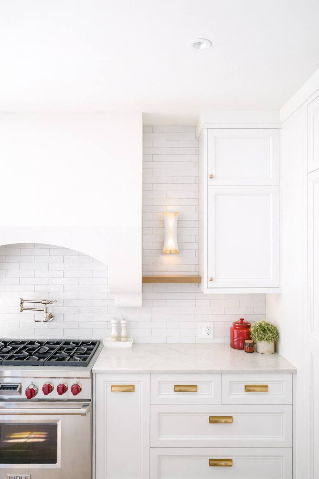 White kitchen with white cabinets, a marble countertop, a stove with red knobs, and decorative items including a red jar, a green plant, and small containers.
