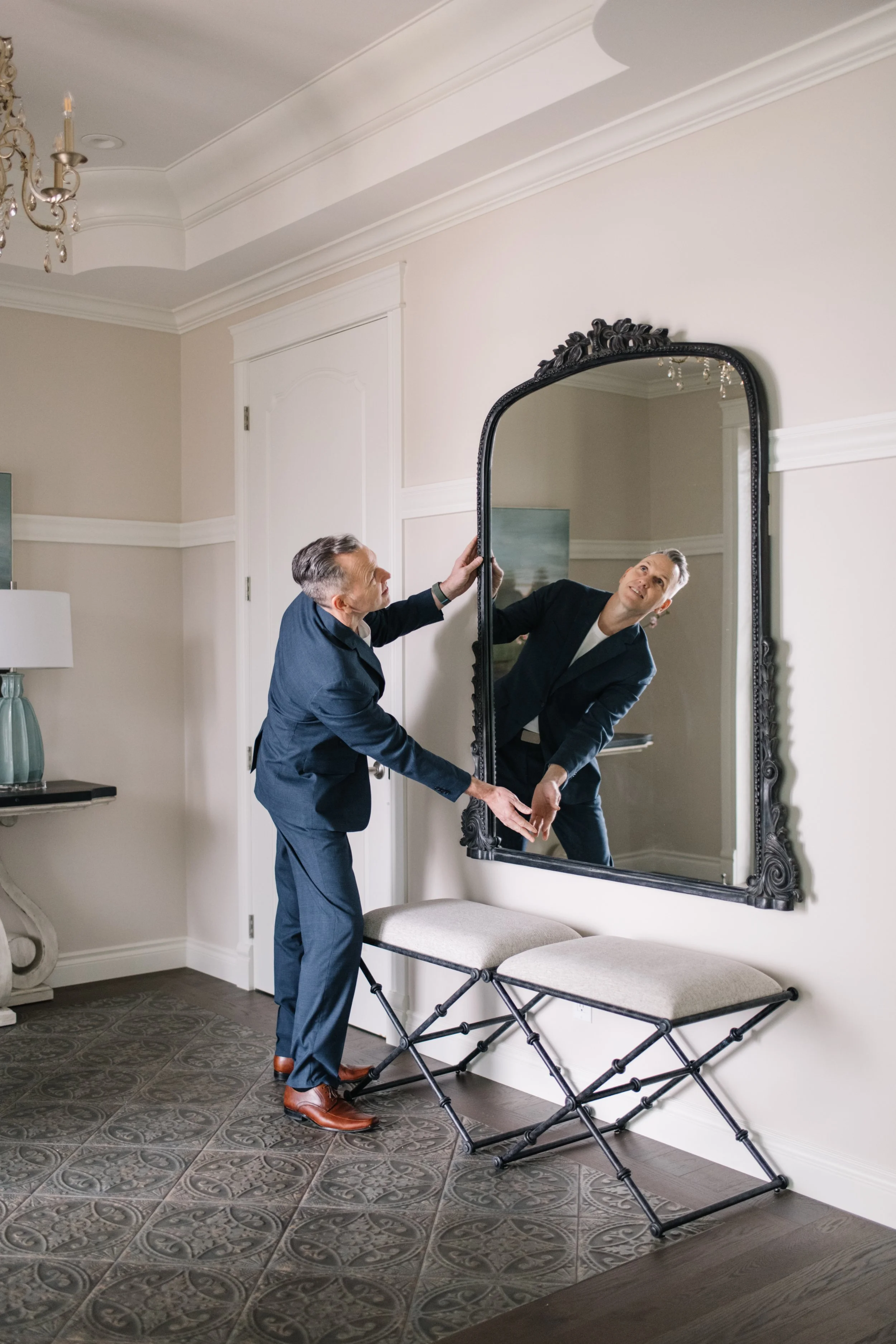 Two men in business suits adjusting a large wall mirror in a decorated room with patterned tile flooring, a chandelier, and a side table with a lamp.