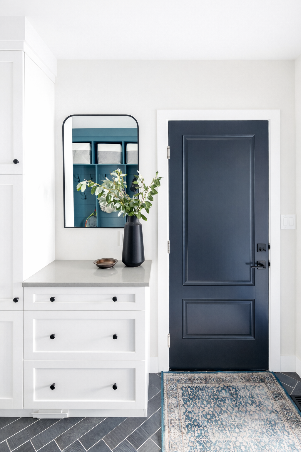 Interior entryway with a black door, white walls, a white cabinet with black knobs, a gray countertop, a black vase with green leaves, a small brown dish, a mirror, and a blue and beige patterned rug.