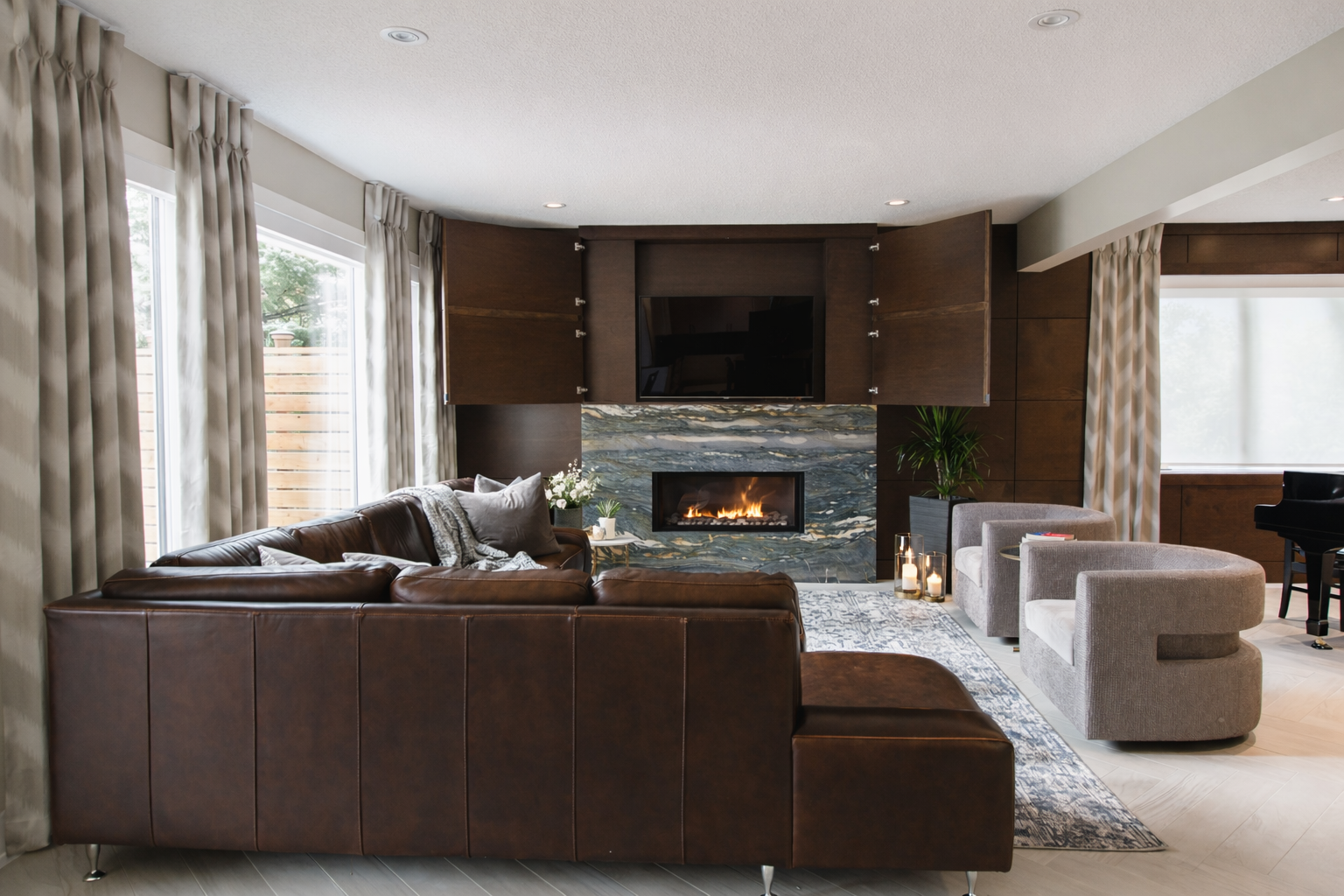 Living room with brown leather sofa, gray chairs, a modern fireplace with a marble surround, a wall-mounted TV, large windows with curtains, and candles.