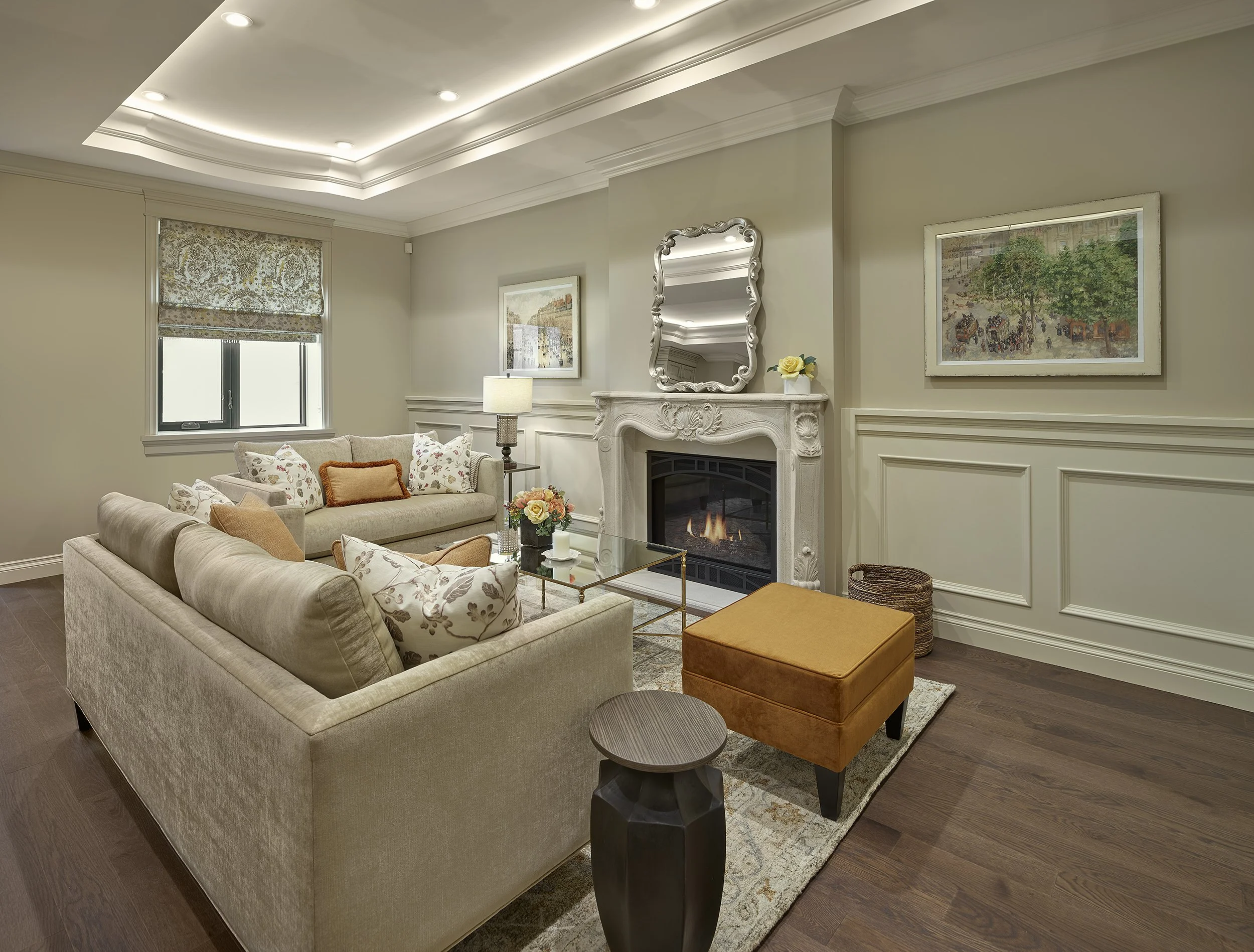 Living room with beige sofas, a fireplace, framed artwork, a mirror above the fireplace, a window with a patterned blinds, a small side table, an orange ottoman, a glass coffee table with flowers, and wooden flooring.