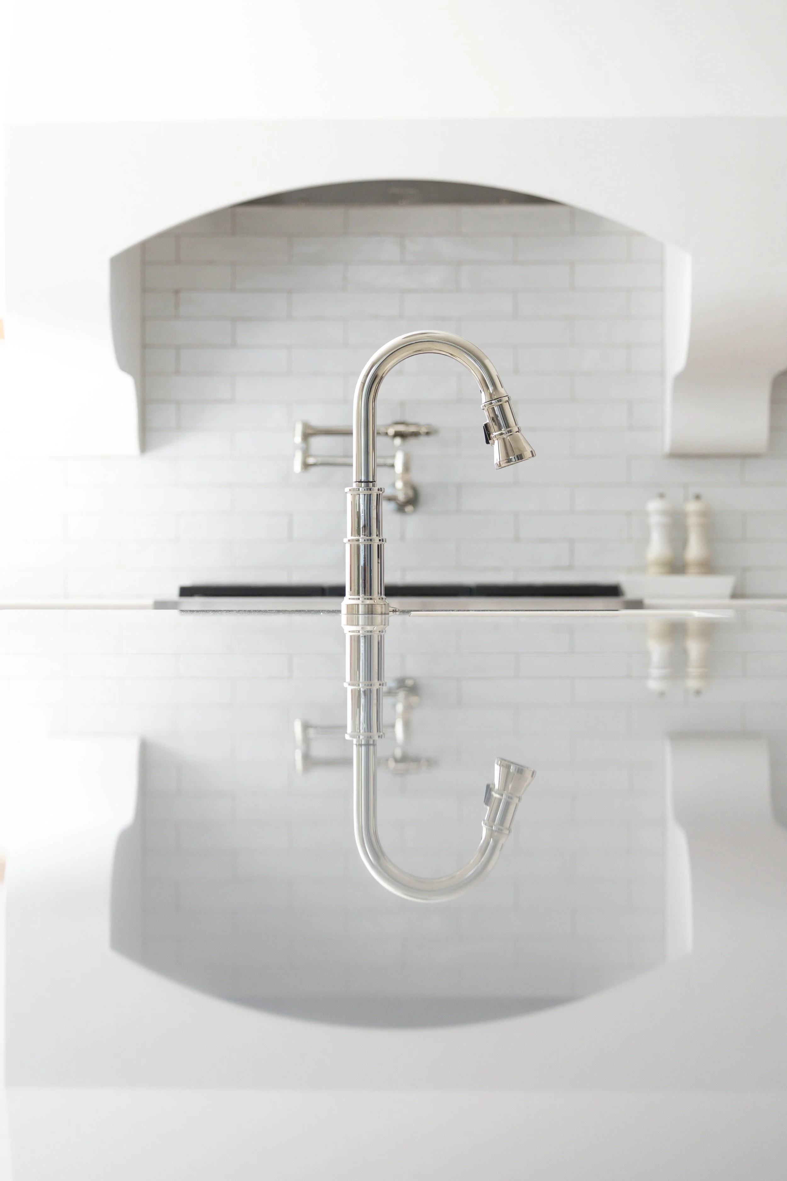 Close-up of a sleek stainless steel kitchen faucet reflected on a white countertop with white brick wall in background.