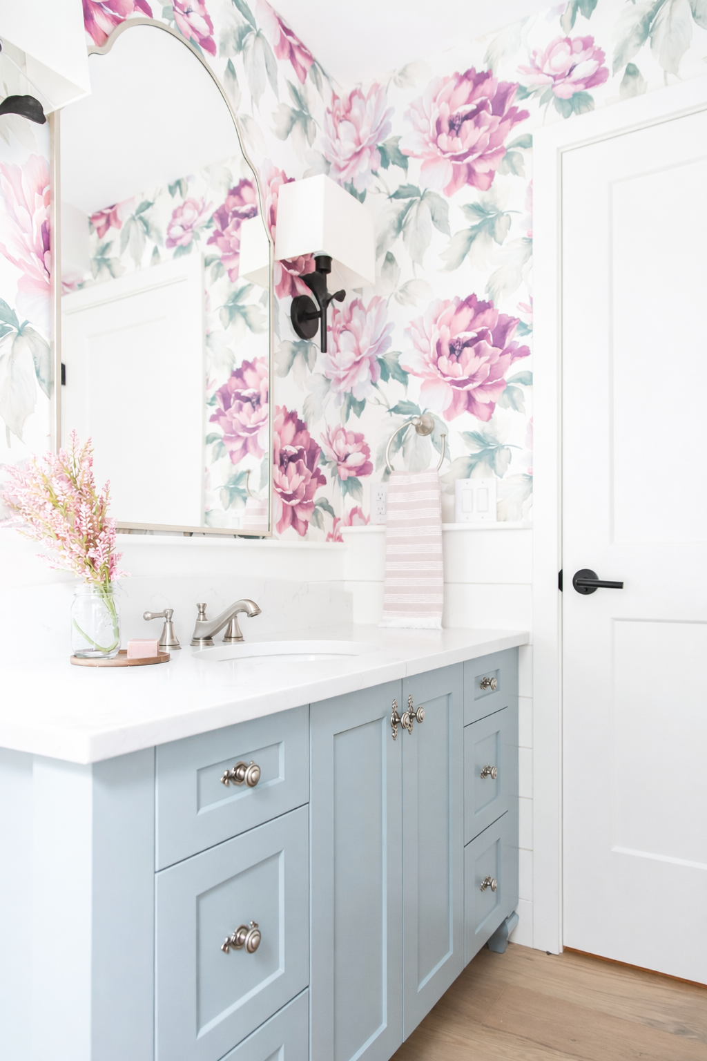 Bathroom with floral wallpaper, blue cabinets, a white countertop, a mirror, a wall-mounted light, a towel ring with a pink and white striped towel, and a vase with pink flowers.