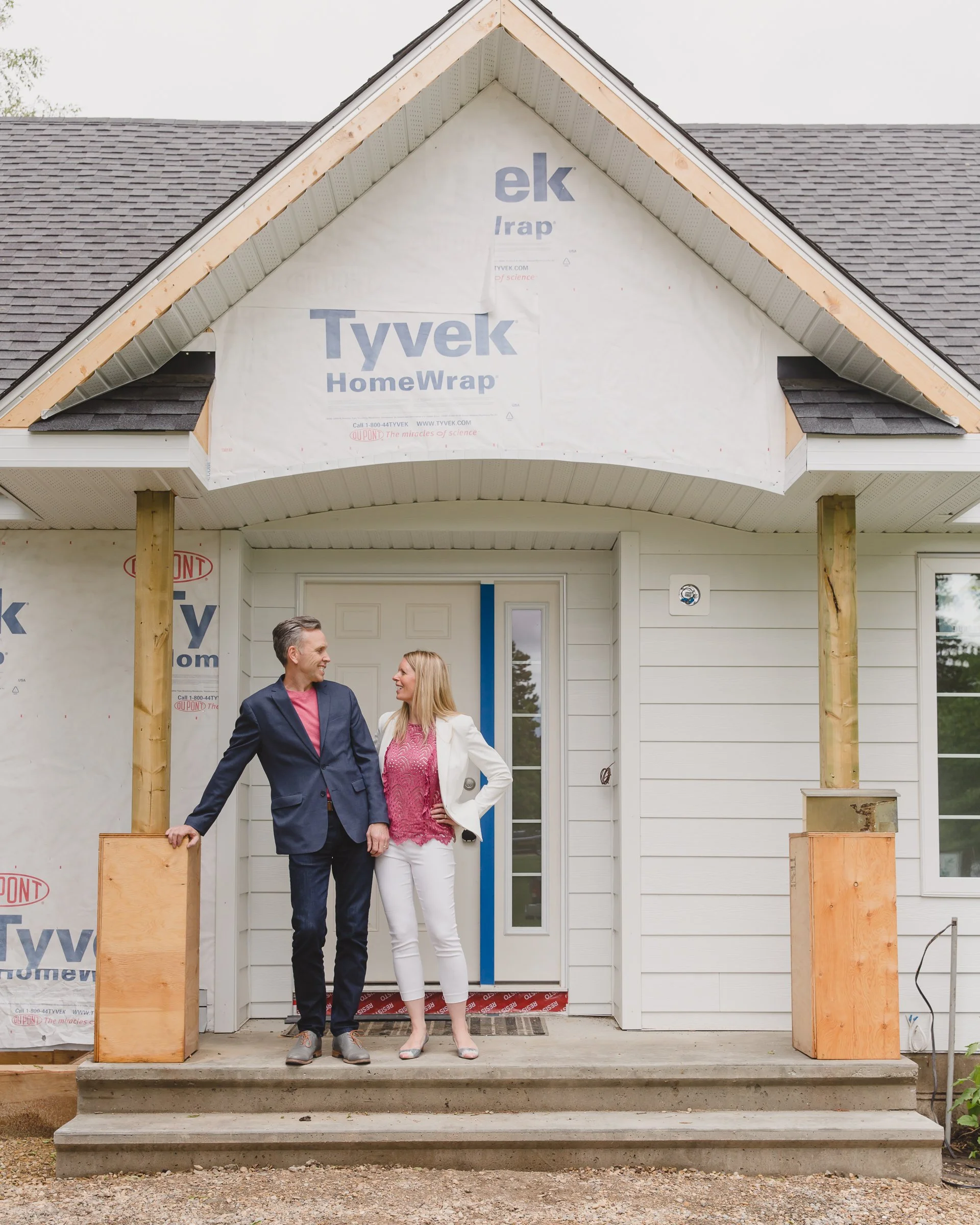 A man and woman in business attire standing on the front porch of a house under construction, smiling and looking at each other.