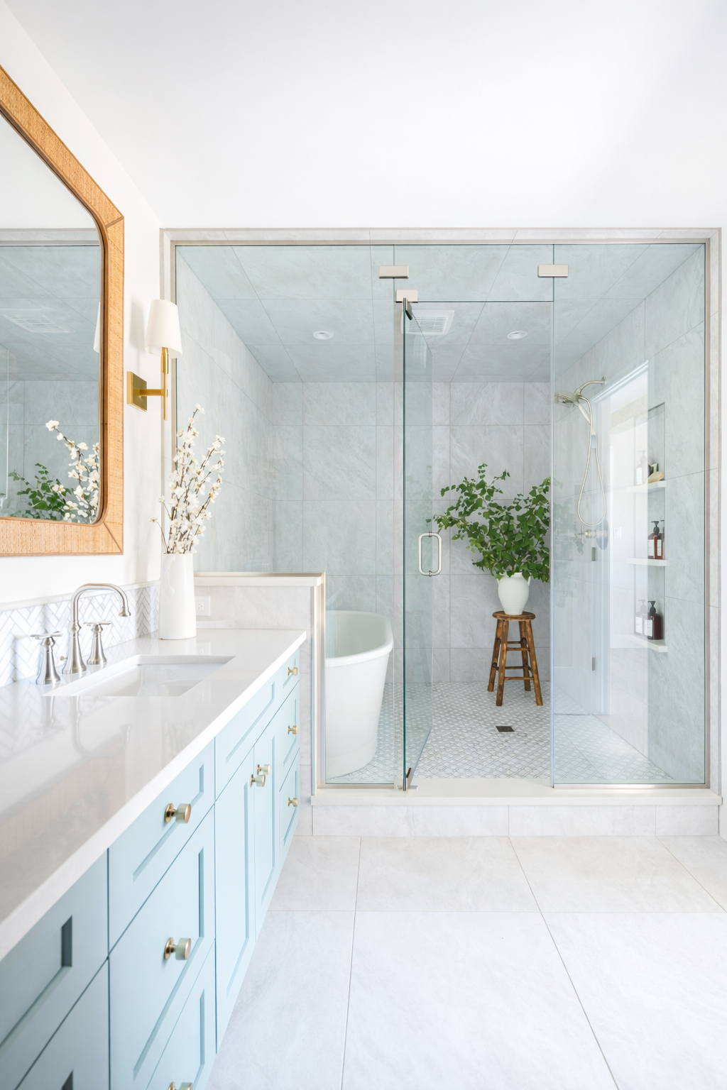 Modern bathroom with white and light blue color scheme, featuring a large mirror, vanity with gold hardware, and a walk-in shower with a glass enclosure, a potted plant, and built-in shelves.
