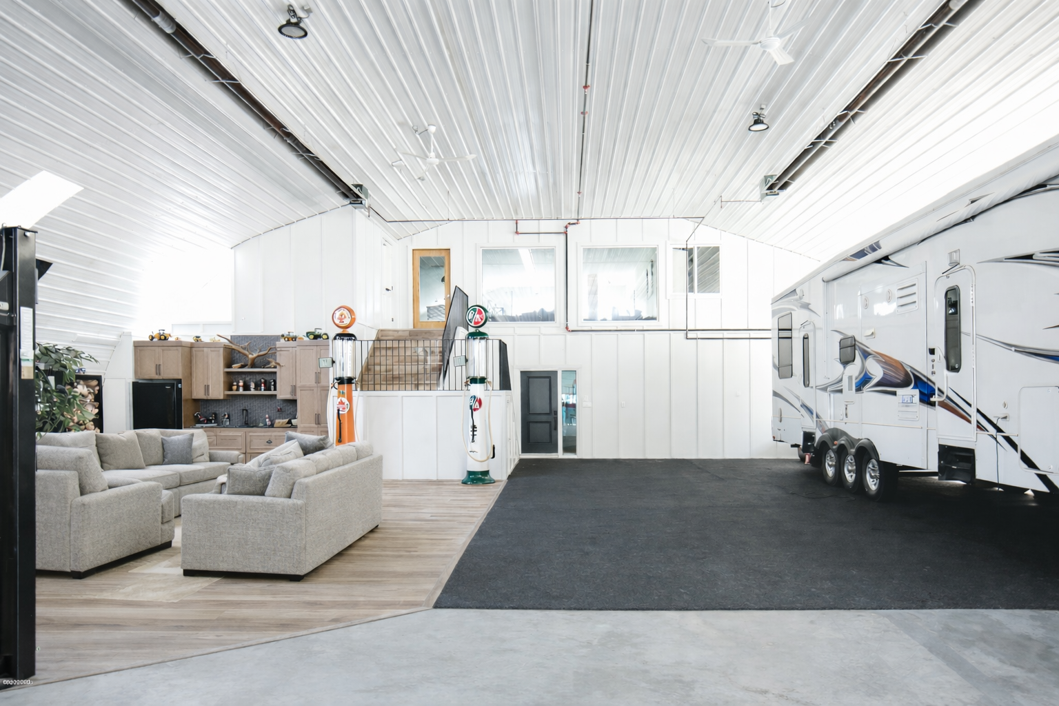 Indoor space with a living area and a large white recreational vehicle (RV). The living area has a beige sofa and a wooden cabinet with shelves. There are vintage gas pump decorations near the stairs. The floor combines wood and carpet, and there is a high, white, vaulted ceiling with lights and ceiling fans.