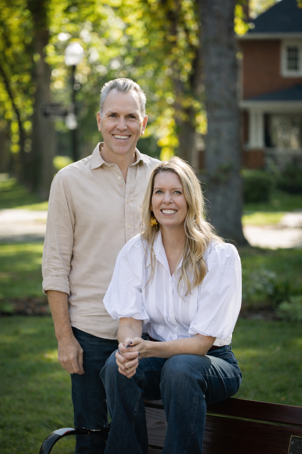 A smiling man standing behind a smiling woman who is sitting on a park bench. They are outdoors with green trees and houses in the background.