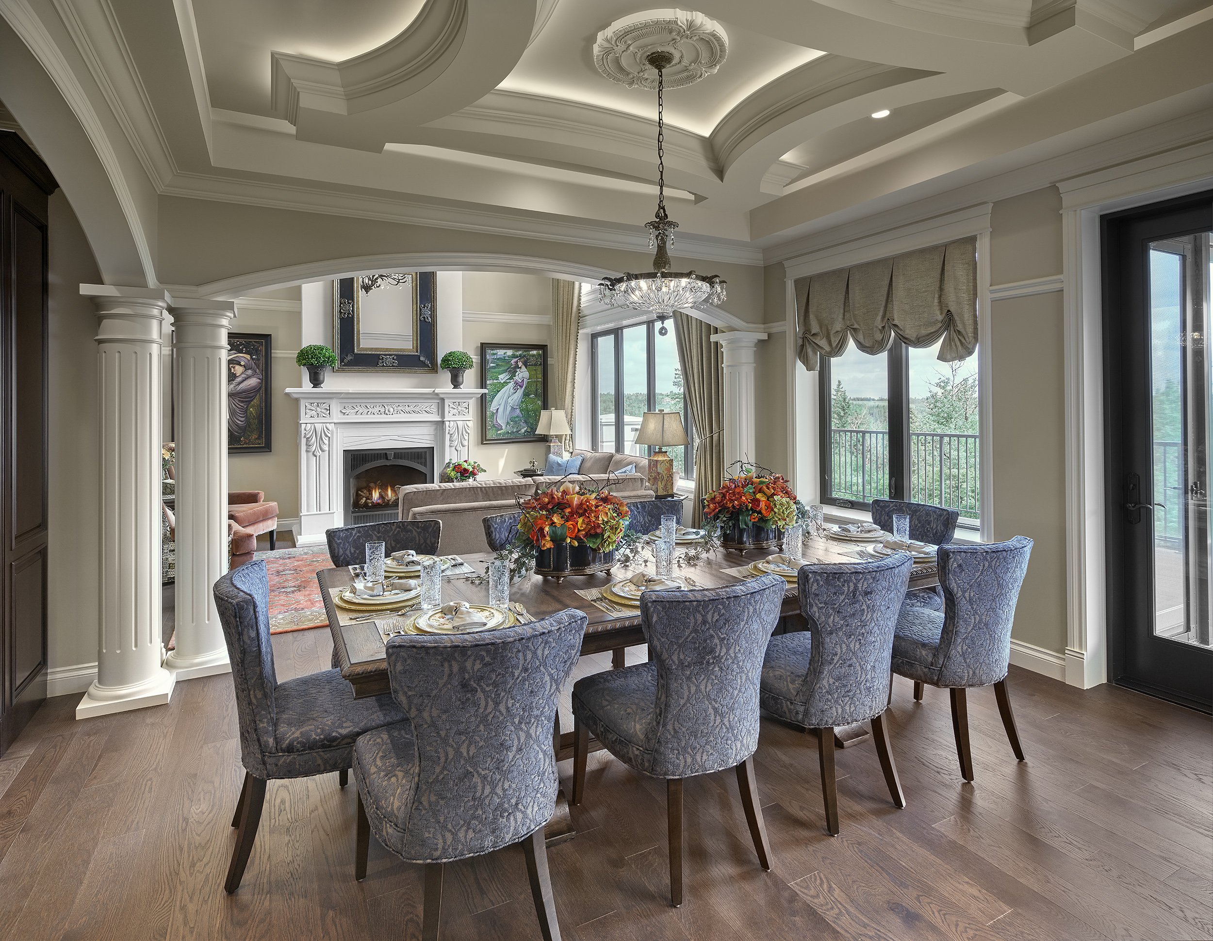 Elegant dining room with wooden table, blue patterned chairs, floral centerpieces, fireplace, artwork, large window, and chandelier.