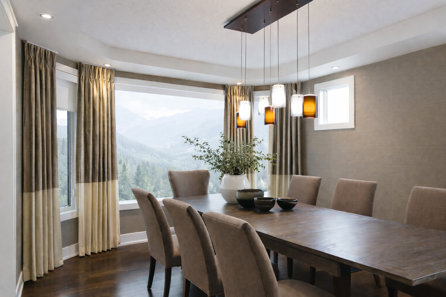A dining room with a large wooden table, beige upholstered chairs, a large window with mountain views, beige curtains, decorative bowls and a plant in a white vase on the table, and modern hanging pendant lights.