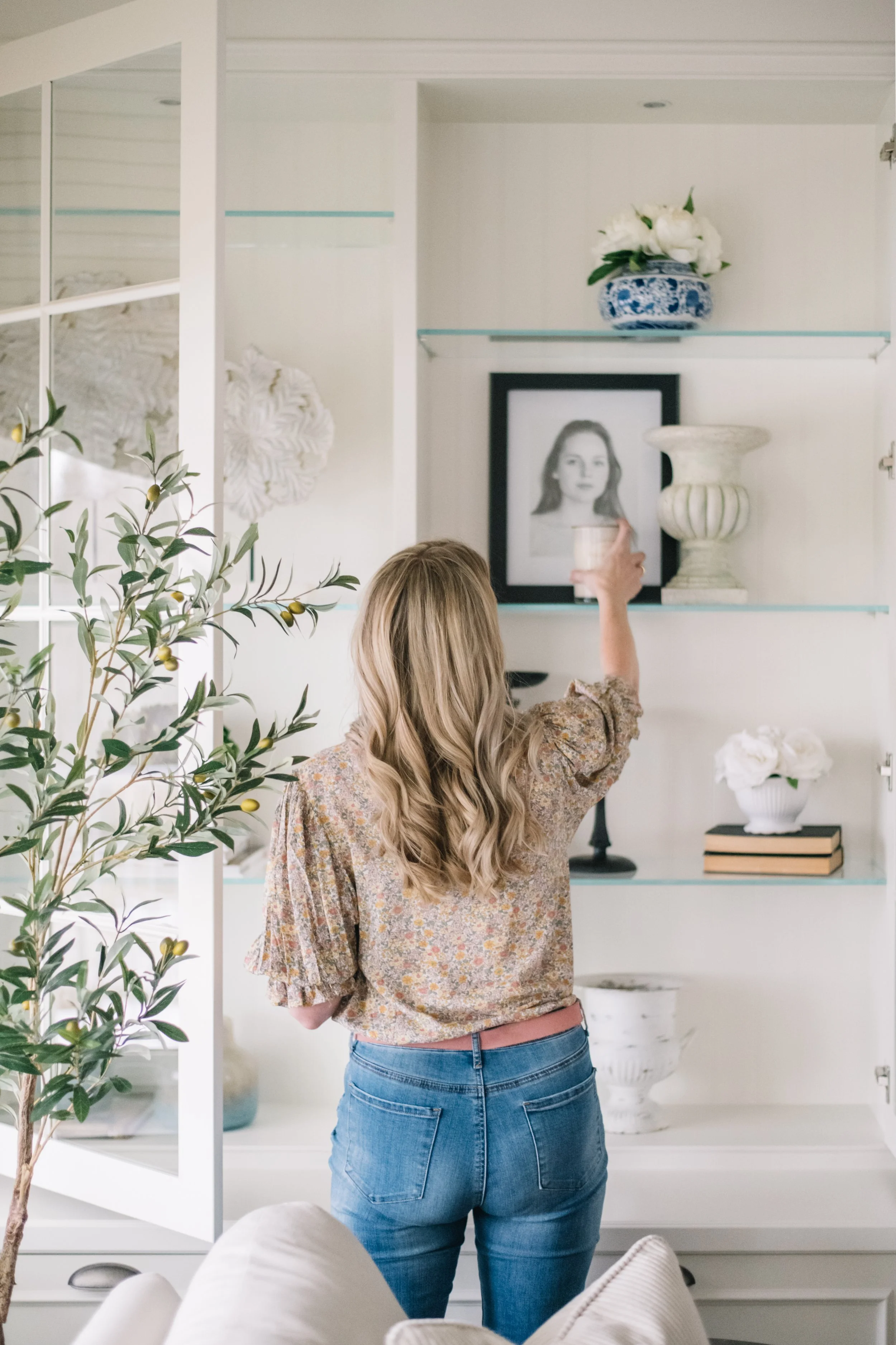 Woman with long blonde hair, wearing a floral blouse and jeans, arranging or adjusting a candle on a white display shelf with decorative items and framed photo.