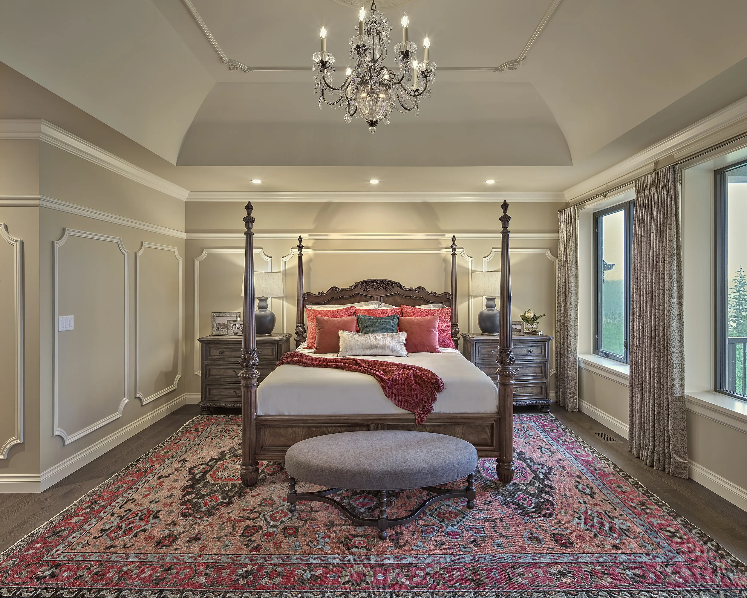 A bedroom with a wooden four-poster bed, red and beige pillows, and a red blanket. There are two nightstands with lamps on each side and a small gray bench at the foot of the bed. The room has cream-colored walls with decorative moldings, a chandelie