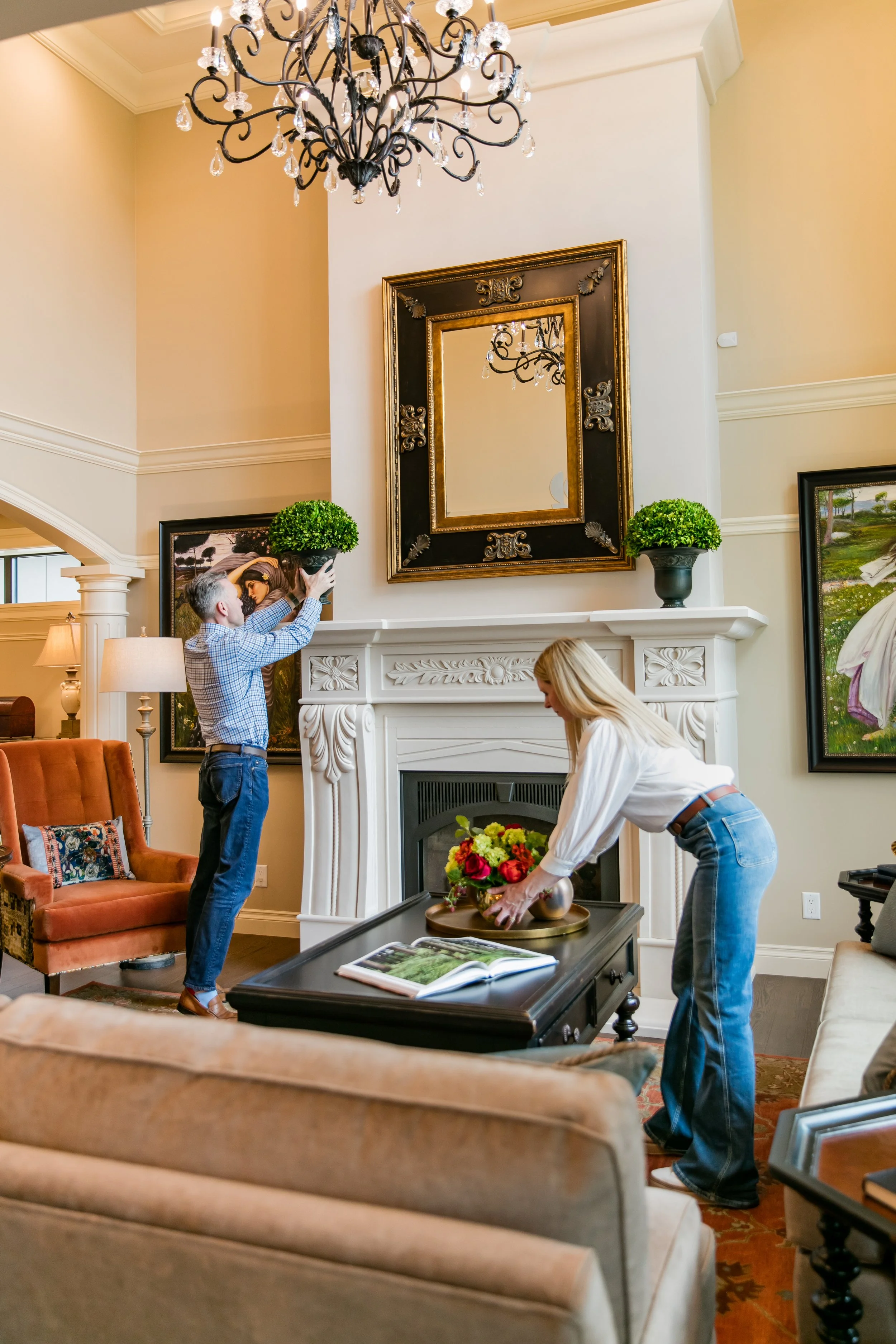 Two women arranging a flower arrangement on a coffee table in a living room with a white fireplace, large mirror, and artwork, while a man puts a potted plant on the mantel.