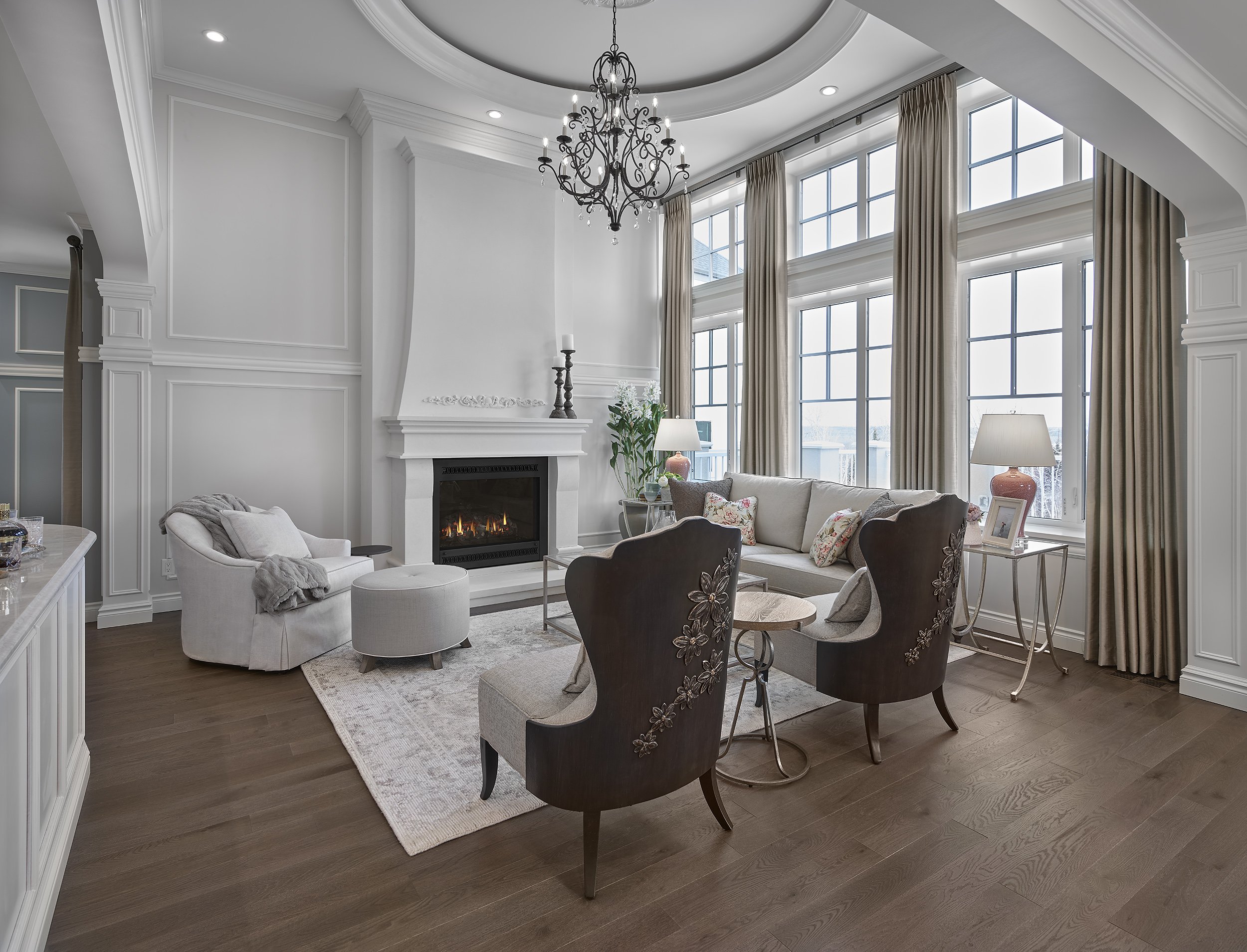 Elegant living room with a white fireplace, large windows with beige curtains, a chandelier, a gray sofa, two dark brown embroidered armchairs, and a small white side table with a picture frame and lamp.