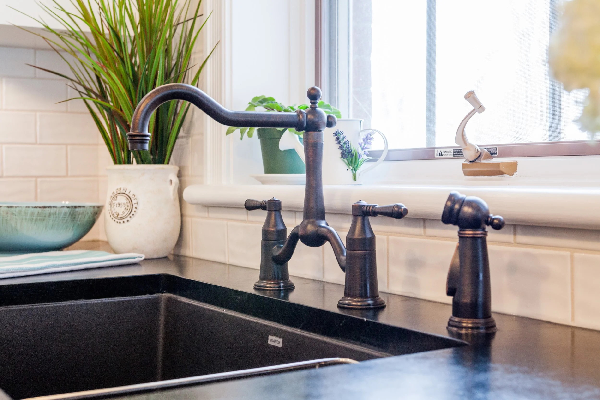 Kitchen sink with a black countertop, bronze faucet, and a window above. There are potted plants and a bowl on the countertop.