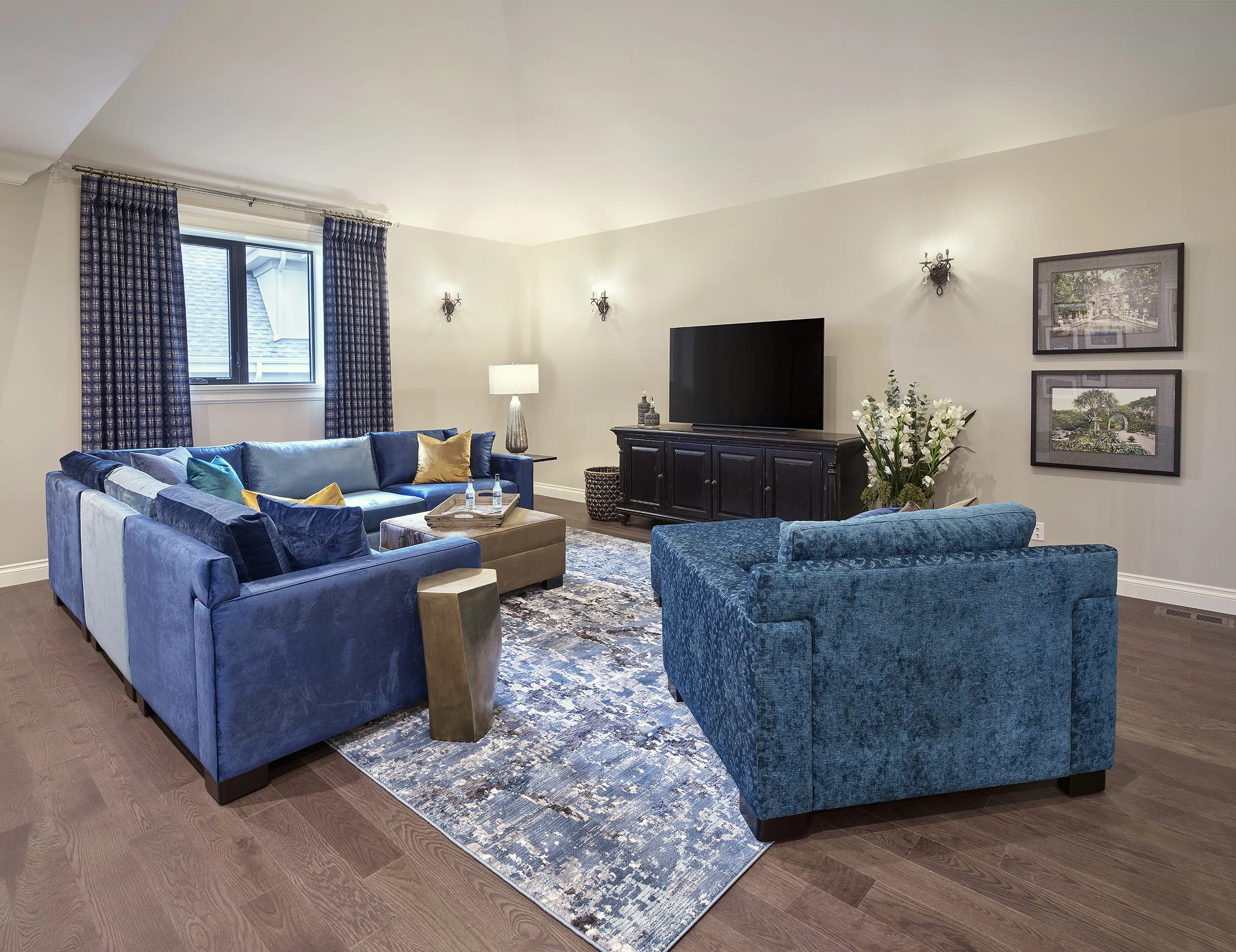 Living room with blue and beige sofas, a TV on a dark wooden stand, framed pictures on the wall, a window with blue curtains, a floral arrangement, and a patterned area rug.