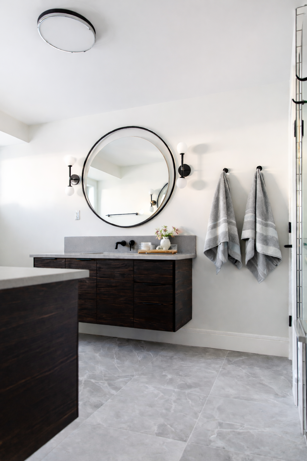 Modern bathroom with dark wood vanity, large round mirror, gray towels on wall, black light fixtures, white walls, and marble-look floor tiles.