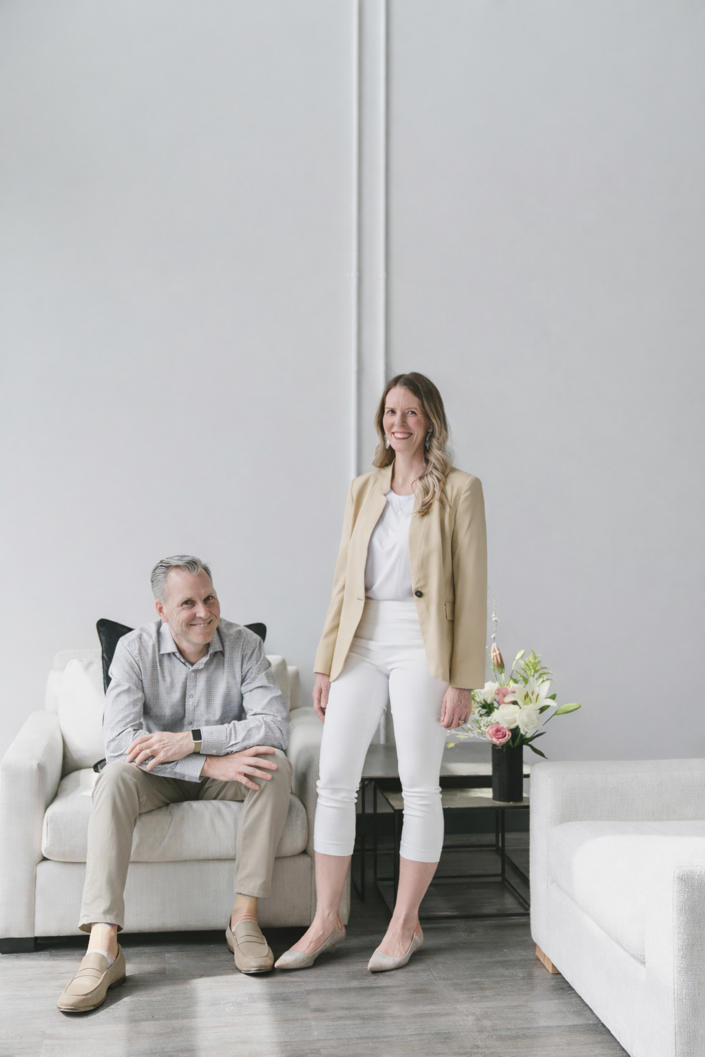 A man seated on a white sofa, smiling, with a smartwatch on his wrist, and a woman standing beside him, smiling, in a modern room with a white sofa, flowers, and minimalist decor.