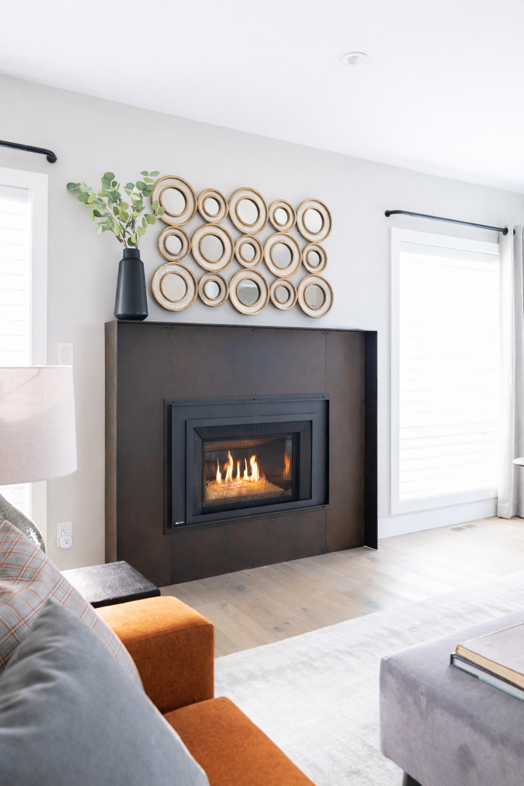 Living room with a modern black fireplace, decorative wooden circles above it, a black vase with eucalyptus on top, and a window with white blinds.