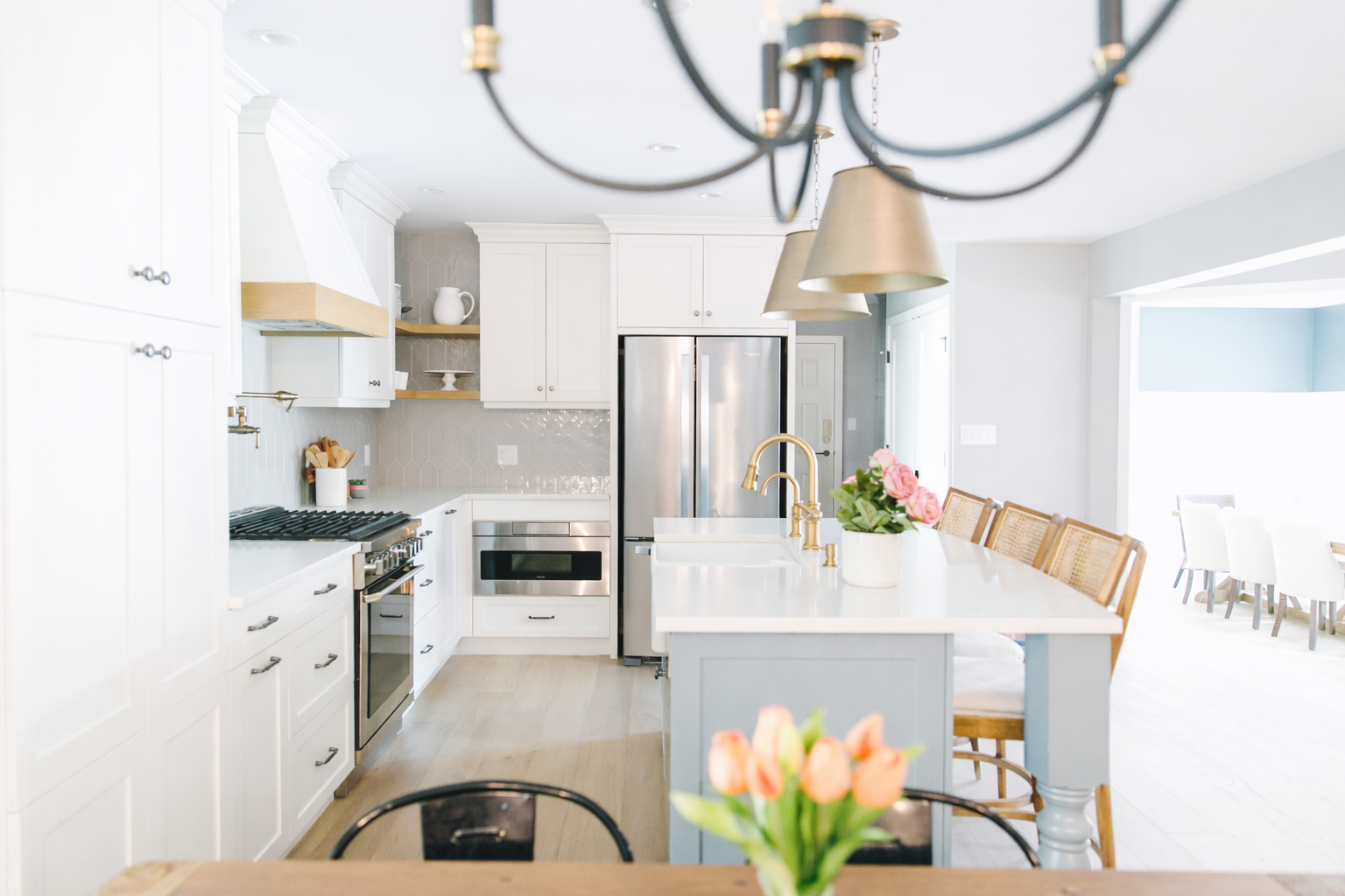 Bright modern kitchen with white cabinets, stainless steel appliances, a central island with a sink, and a chandelier overhead. There are pink flowers in a white vase on the island.