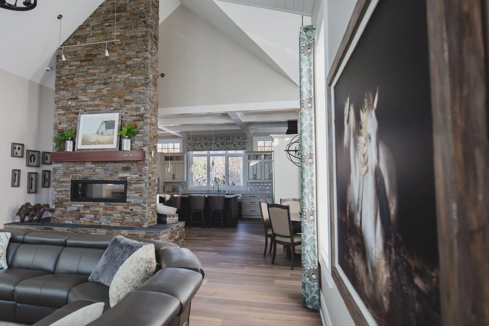 Living room with a stone fireplace, leather sofa, and view into the kitchen and dining area with large windows, patterned curtains, and modern decor.
