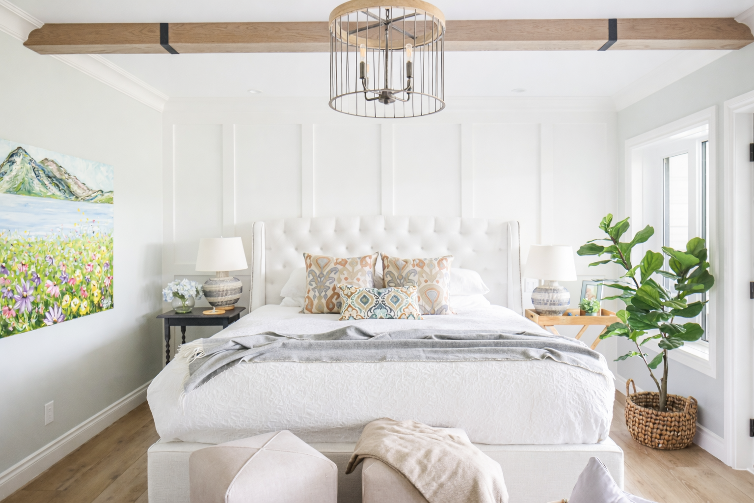 Bright bedroom with a large white tufted headboard, colorful patterned pillows, and white bedding. Two bedside tables with lamps, a large green potted plant in a woven basket, and a landscape painting on the left wall. Overhead light fixture and windows on the right side.