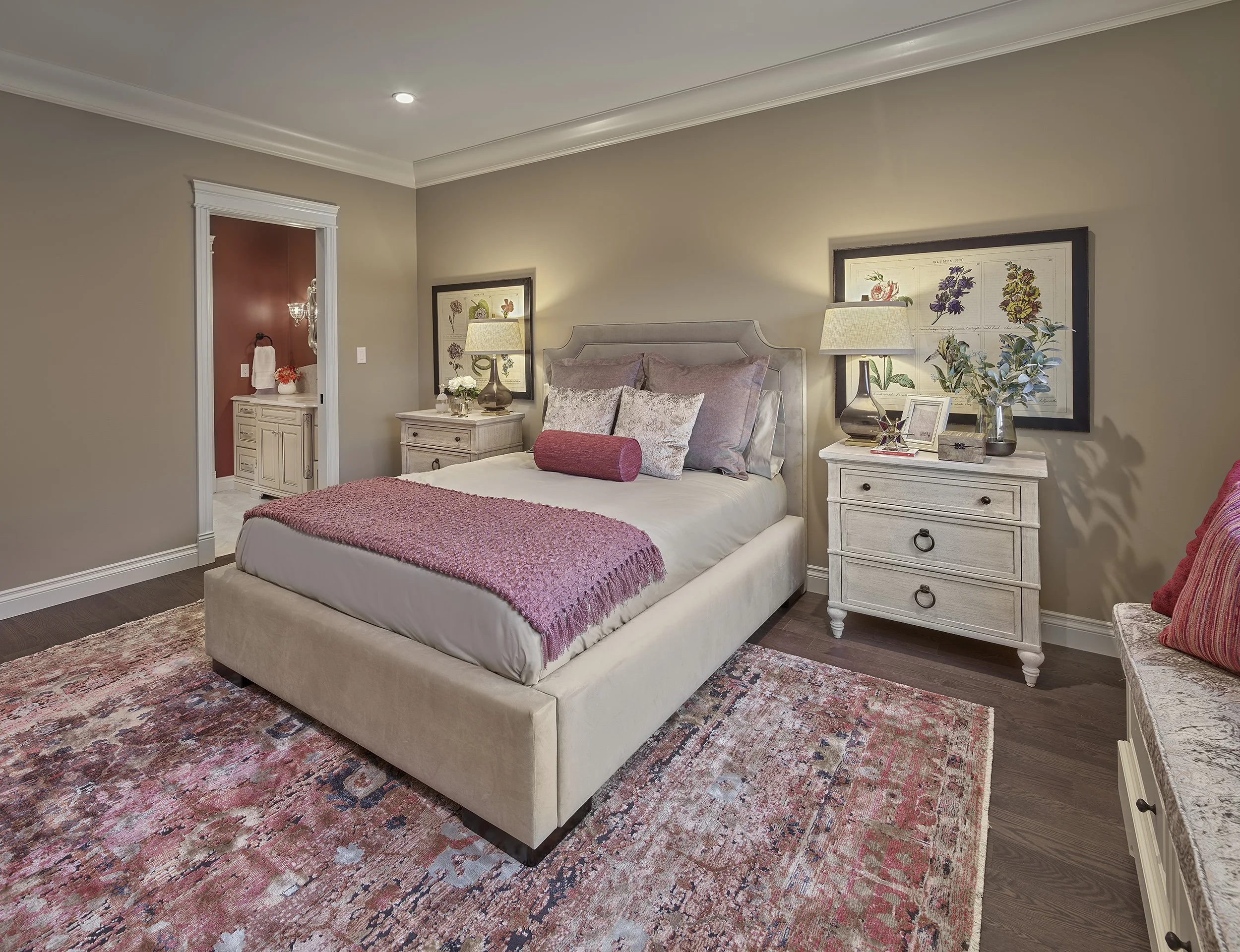 A cozy bedroom with a beige upholstered bed, pink throw, and multiple decorative pillows. Two white nightstands with lamps, framed botanical prints, and plants. A colorful area rug on dark hardwood flooring, with an adjacent bathroom visible through 