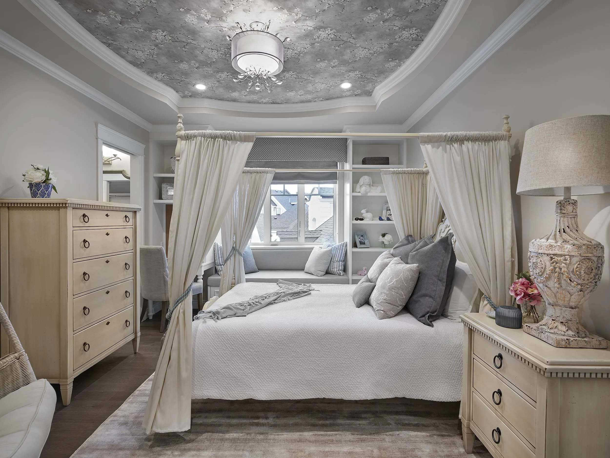 A bedroom with a four-poster bed surrounded by white curtains, a dresser, a nightstand with a lamp and vase, and a window seat with cushions, decorated in neutral tones.