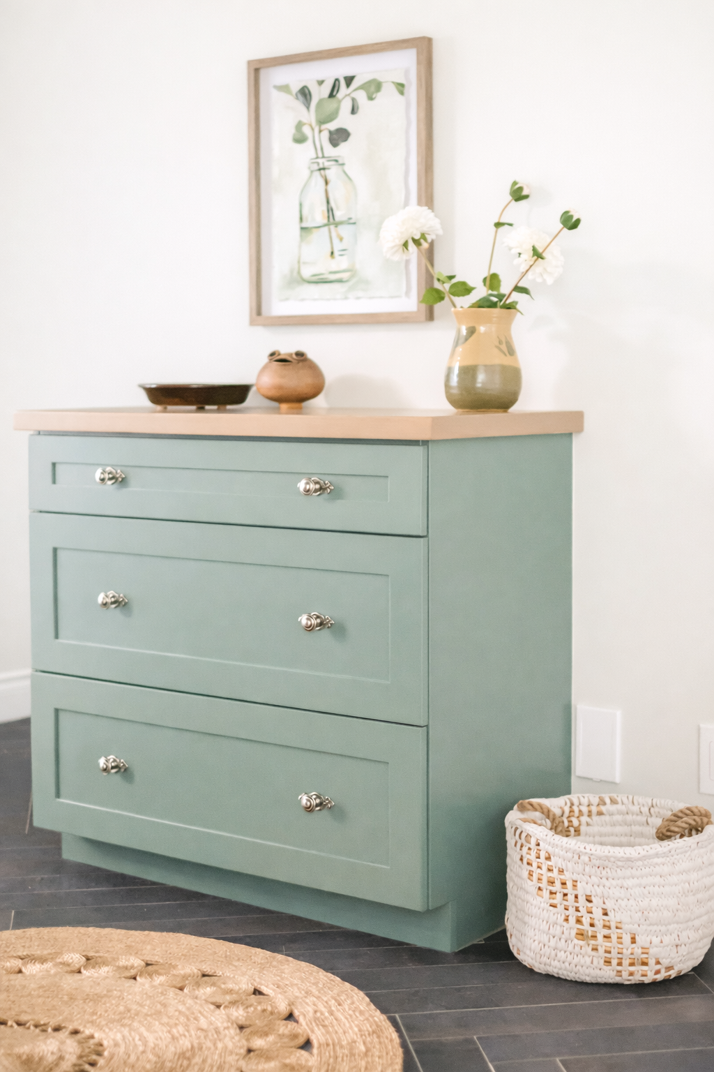 A light green dresser with four drawers, silver knobs, topped with a beige wooden surface. On the surface, there is a small ceramic brown pot, a shallow brown dish, and a ceramic vase with white flowers and green leaves. Above the dresser, a framed w