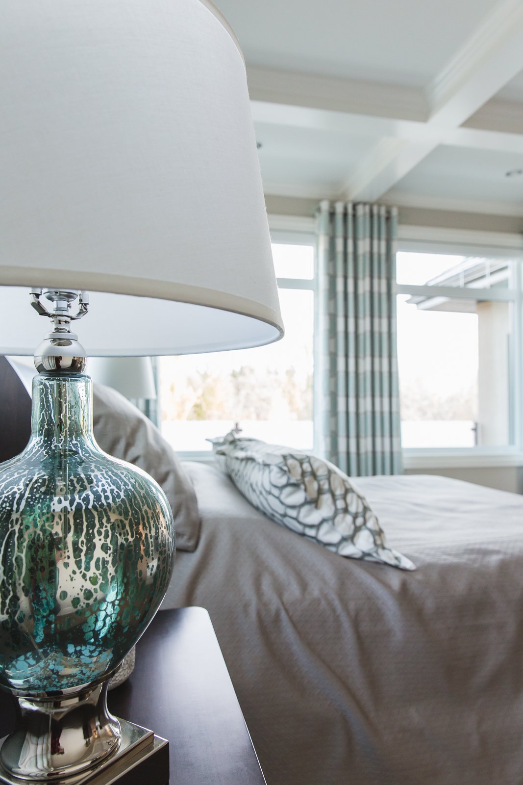 Close-up of a bedside table with a decorative silver and blue glass table lamp, with a bedroom in the background featuring a bed, pillows, large windows, and striped curtains.