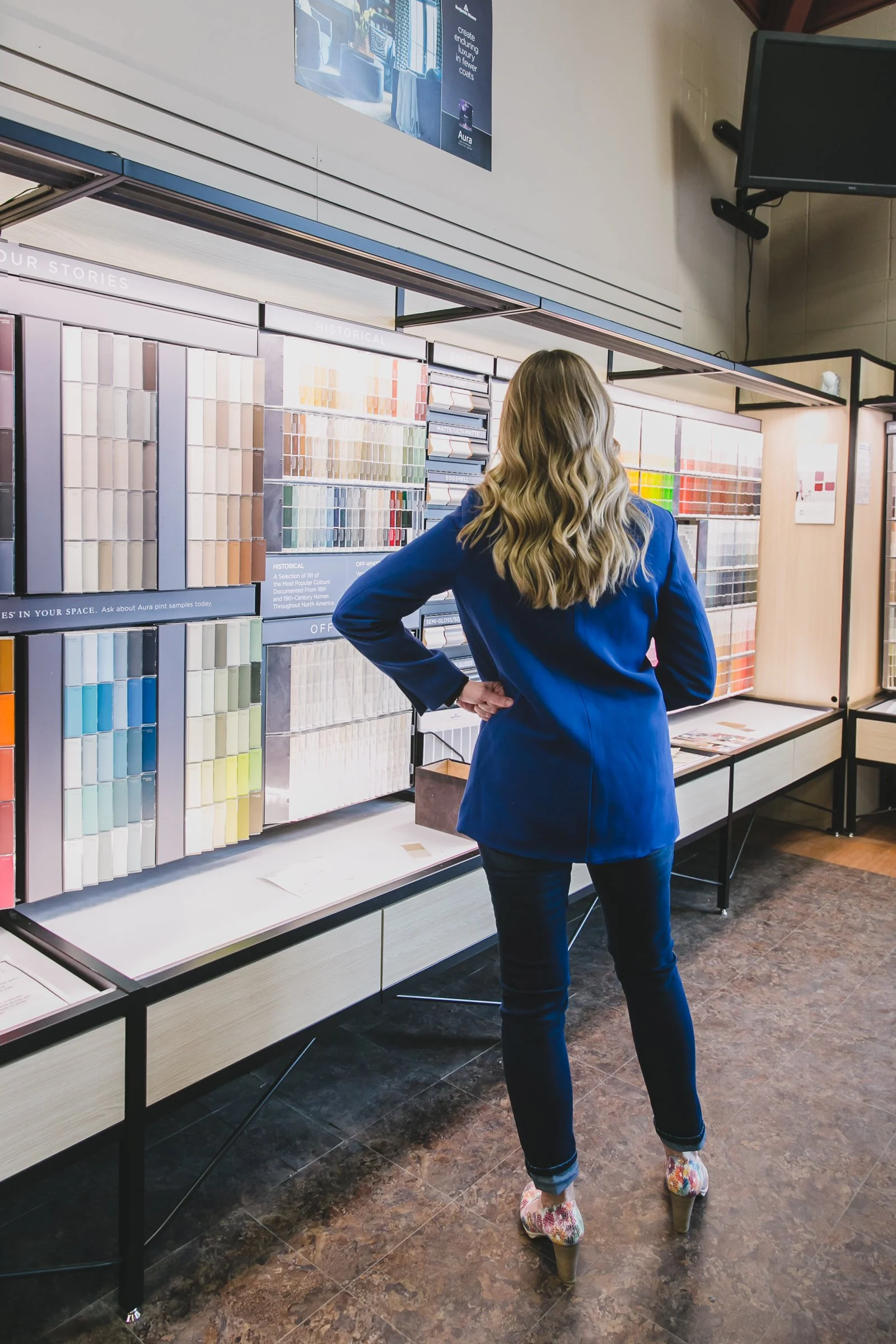 A woman with long, wavy blonde hair, dressed in a blue jacket, dark jeans, and high-heeled floral shoes, is standing in front of a display of paint samples in a store, looking at the color options.