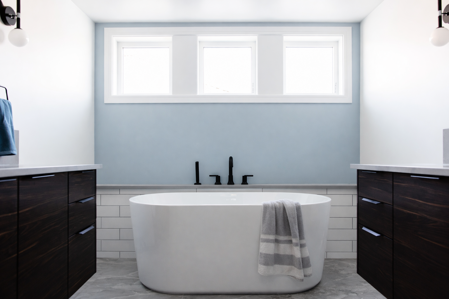 Modern bathroom featuring a white freestanding bathtub with a gray striped towel draped over its edge, dark wooden vanity cabinets on each side, white tiled walls, and a high window letting in natural light.