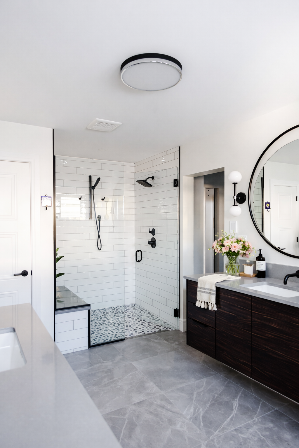 Modern bathroom with a glass-enclosed walk-in shower featuring white subway tiles and black fixtures, a dark wood vanity with a white countertop, a round mirror, a bouquet of flowers, and a contemporary ceiling light.