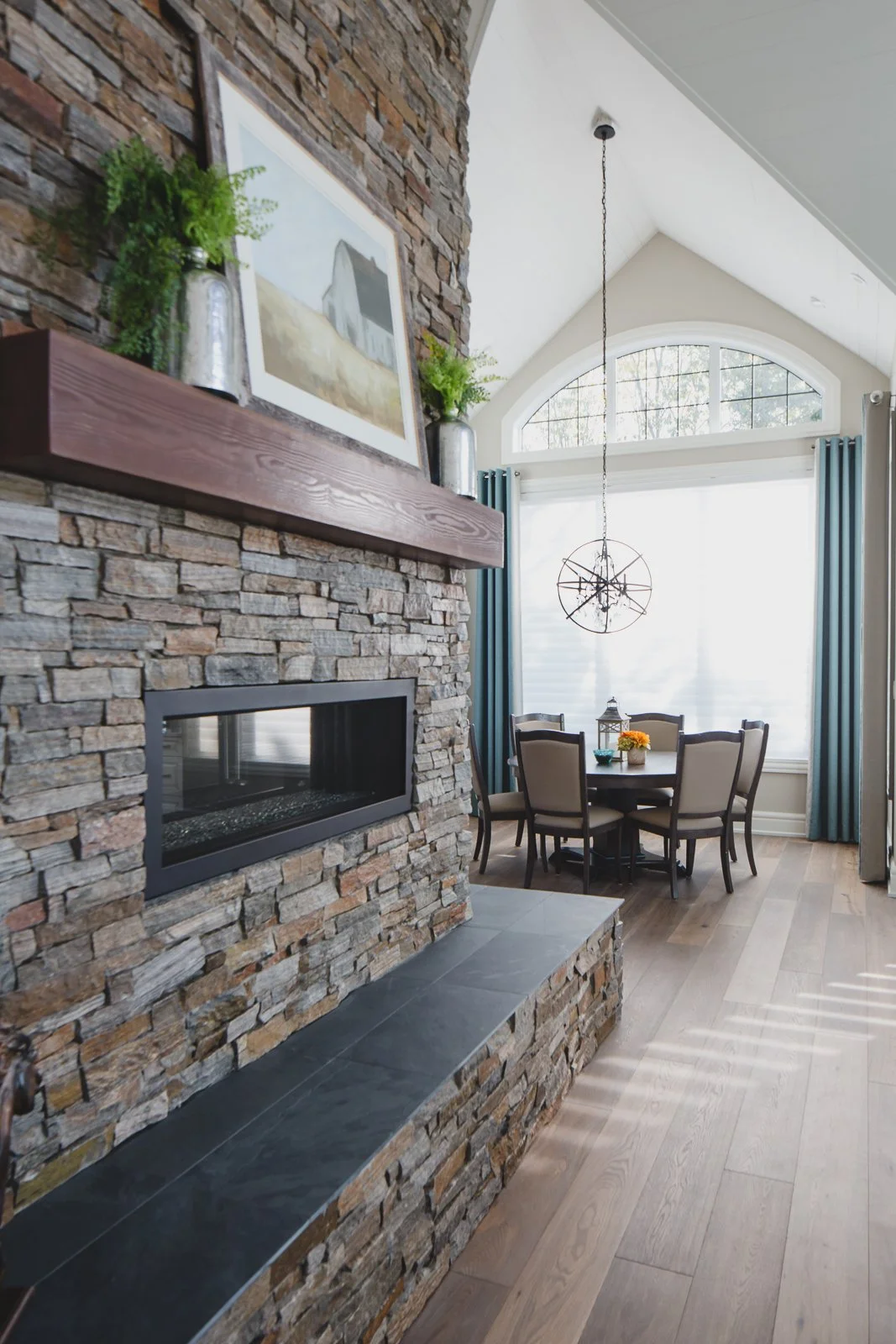 Living room with a stone fireplace, a round dining table with eight chairs, large arched window with curtains, hanging light fixture, and wood flooring.