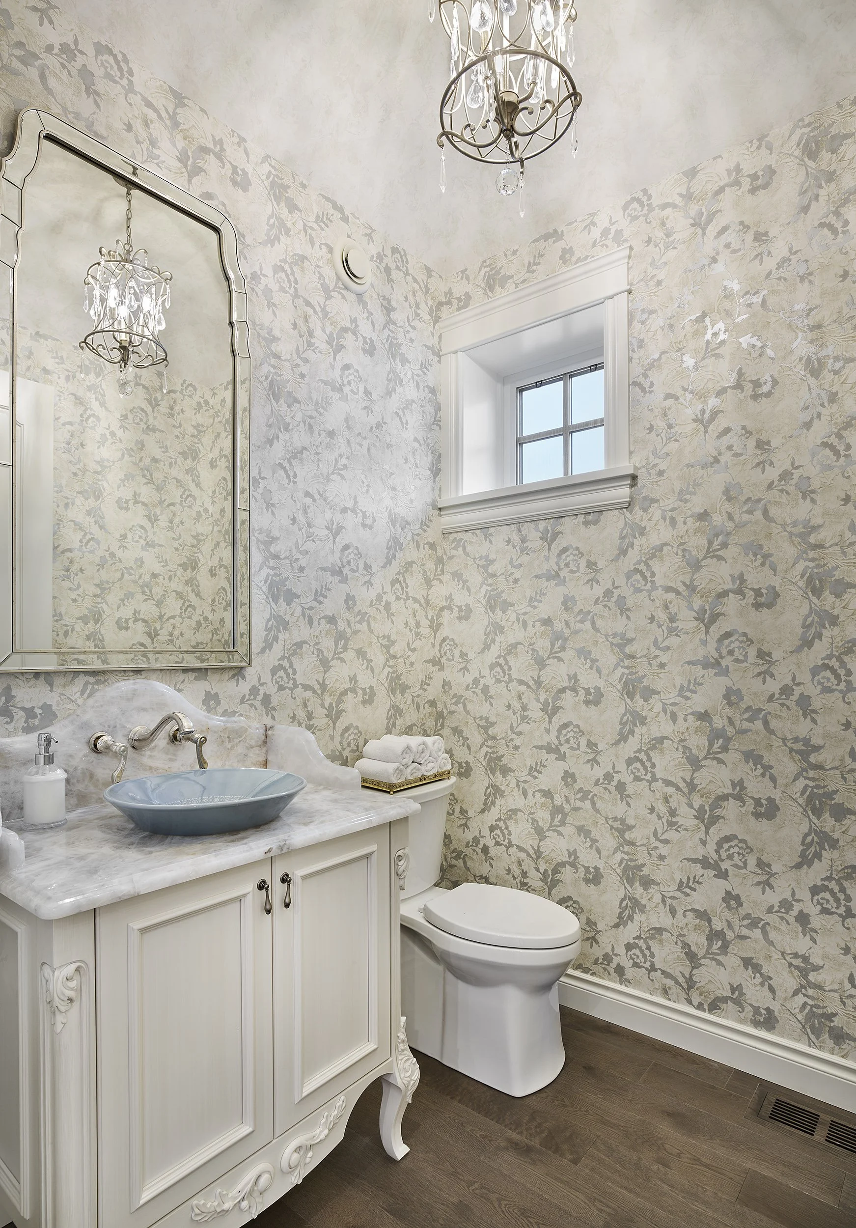 A vintage bathroom with floral-patterned wallpaper, a white wooden cabinet with a marble top, a blue vessel sink, a mirror, a small window, and a chandelier.