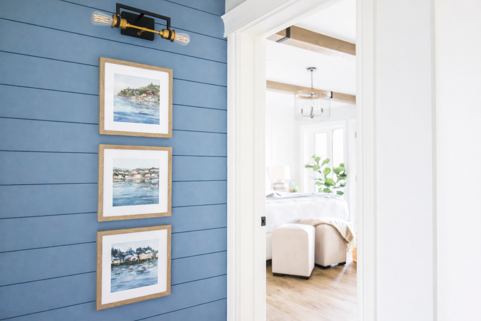Interior view of a house with a blue shiplap wall display and three landscape paintings framed in light wood, seen through an open doorway into a bright living room with white walls, wooden beams, window, and green plant.